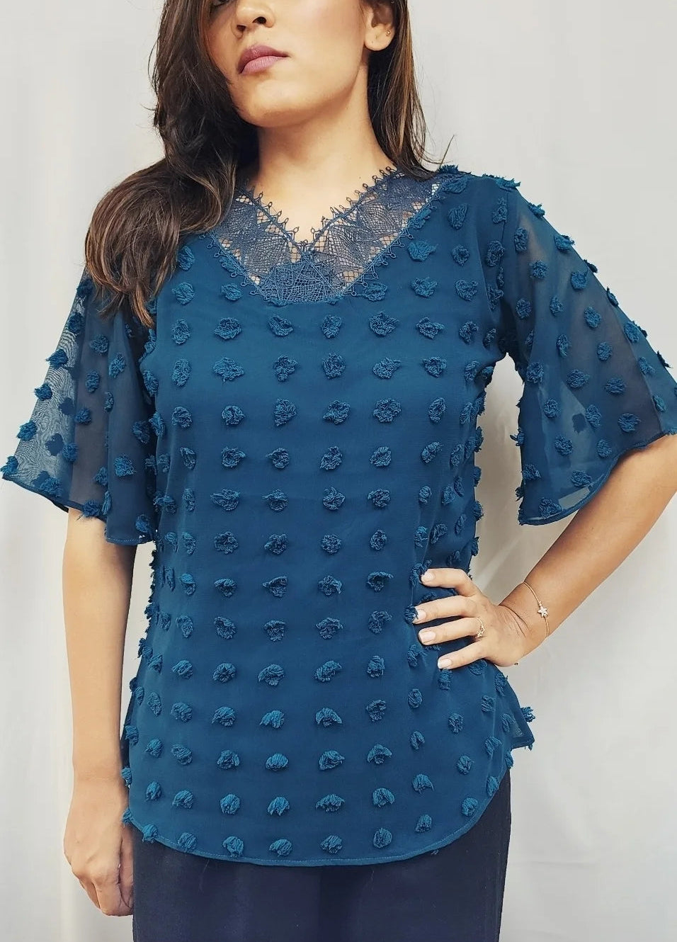 A woman wearing a textured teal blue short-sleeve blouse with a lace V-neckline, standing against a plain light gray background.