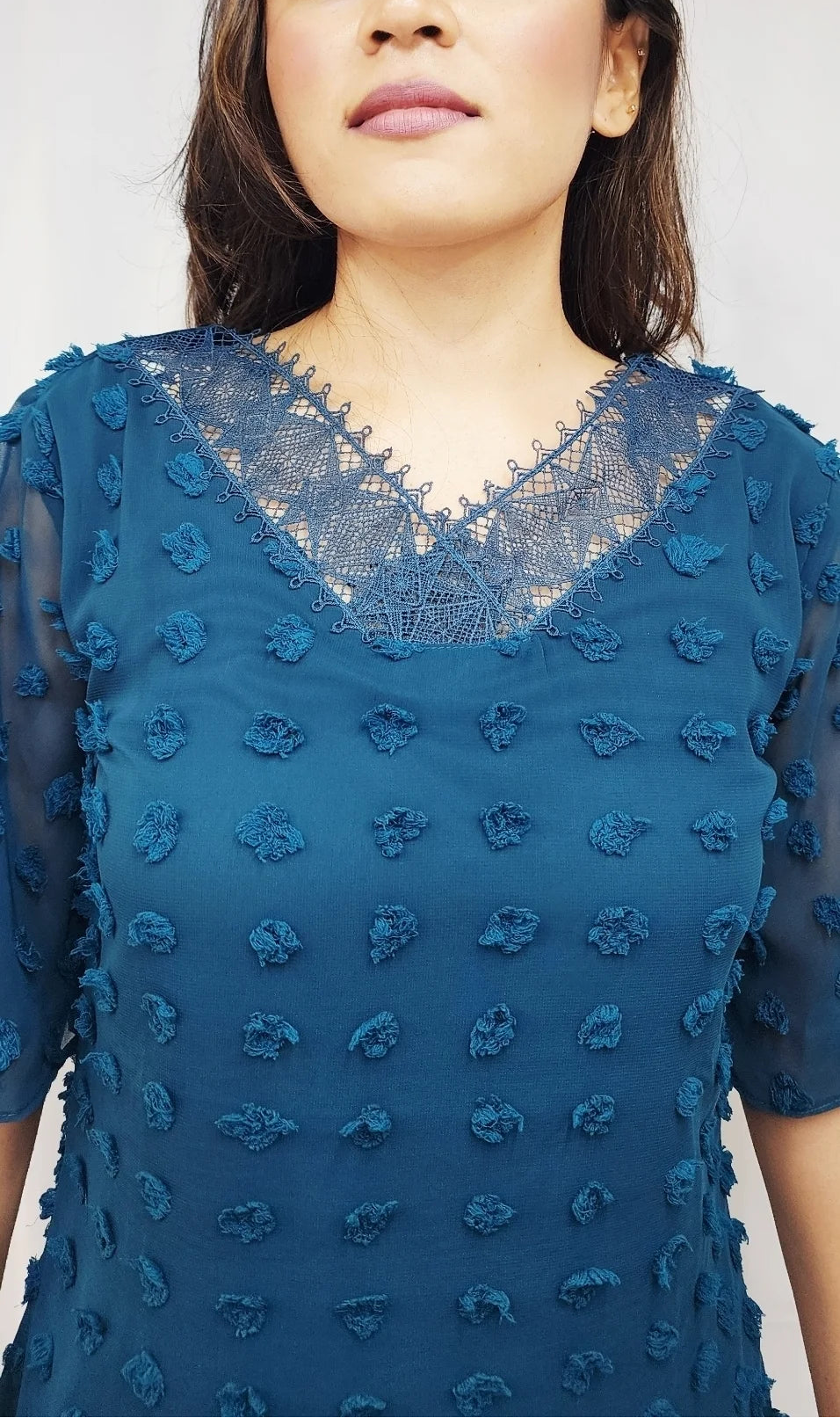 A woman wearing a teal blue textured lace top with short sleeves and a V-neck, shown from the chest up against a plain light background.