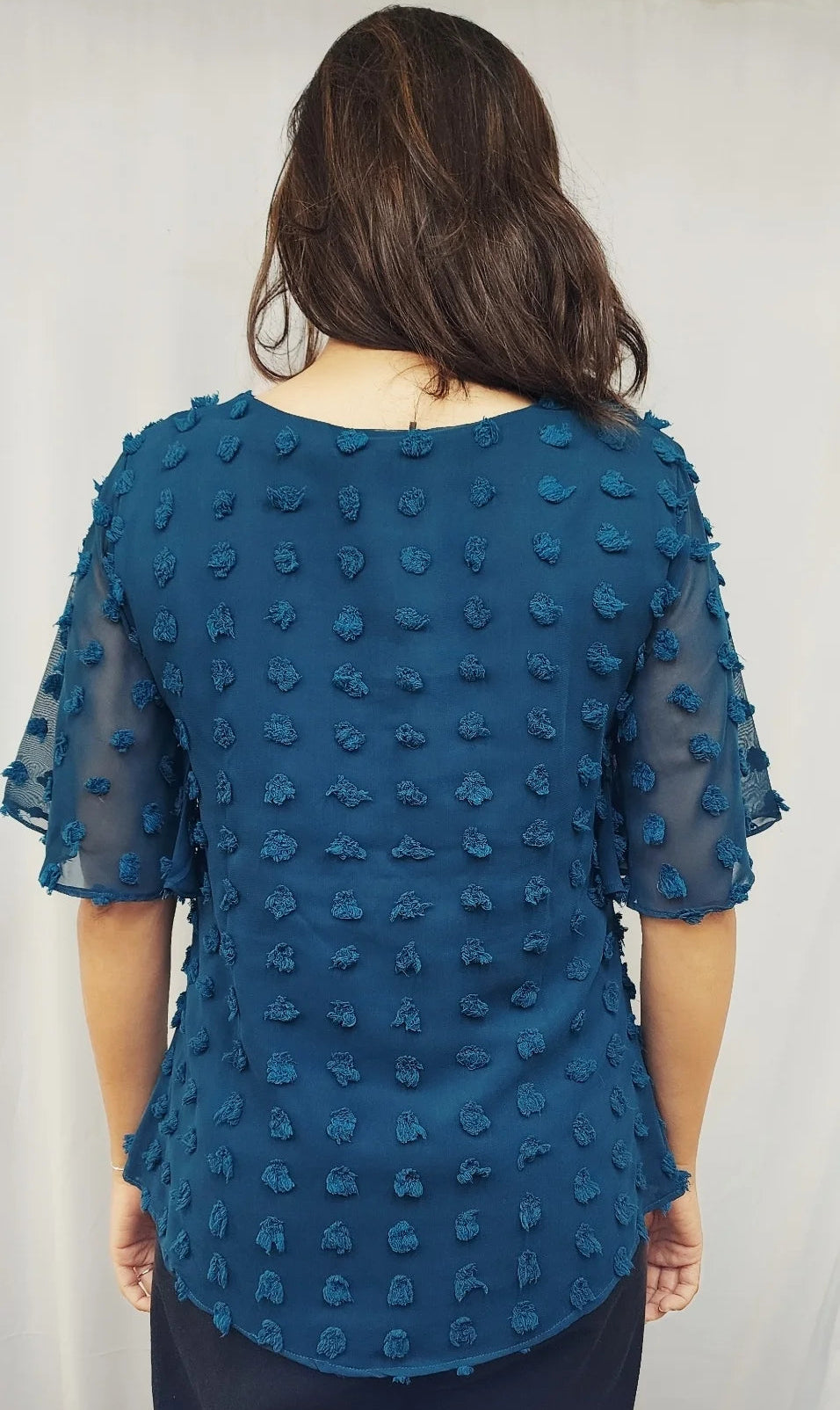A woman viewed from the back wearing a teal blue textured short-sleeve blouse with sheer flutter sleeves and dark bottoms, standing against a white curtain backdrop.
