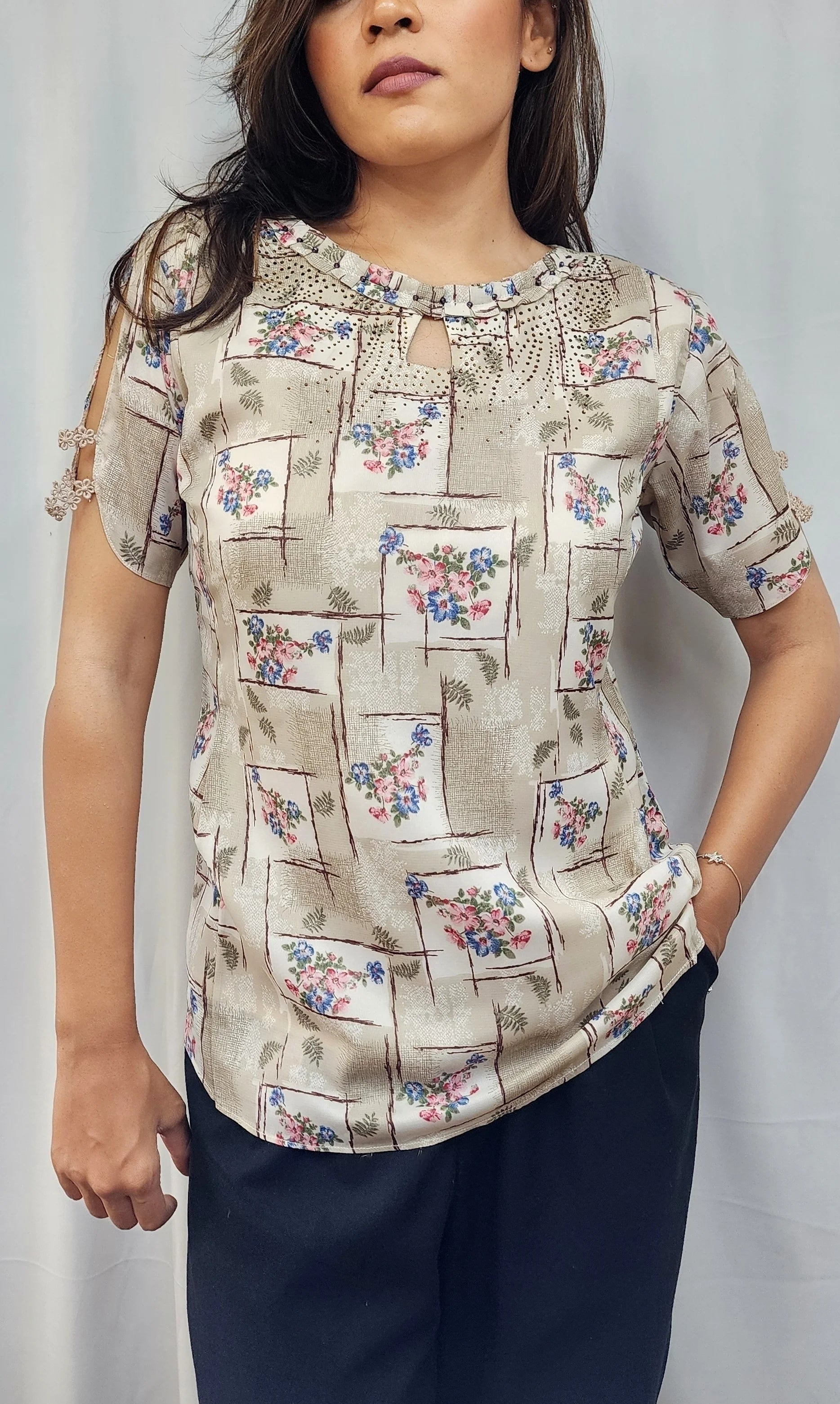 A woman modeling a short-sleeve beige blouse with a floral and geometric patchwork print, paired with dark navy pants, standing against a light gray curtain backdrop.