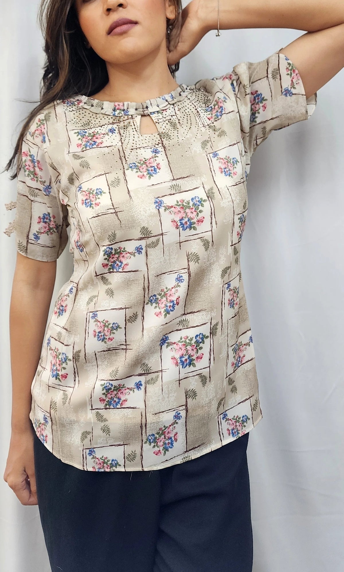 A woman modeling a beige short-sleeve top with a multicolor floral and geometric block print, paired with dark blue pants, standing against a white curtain backdrop.