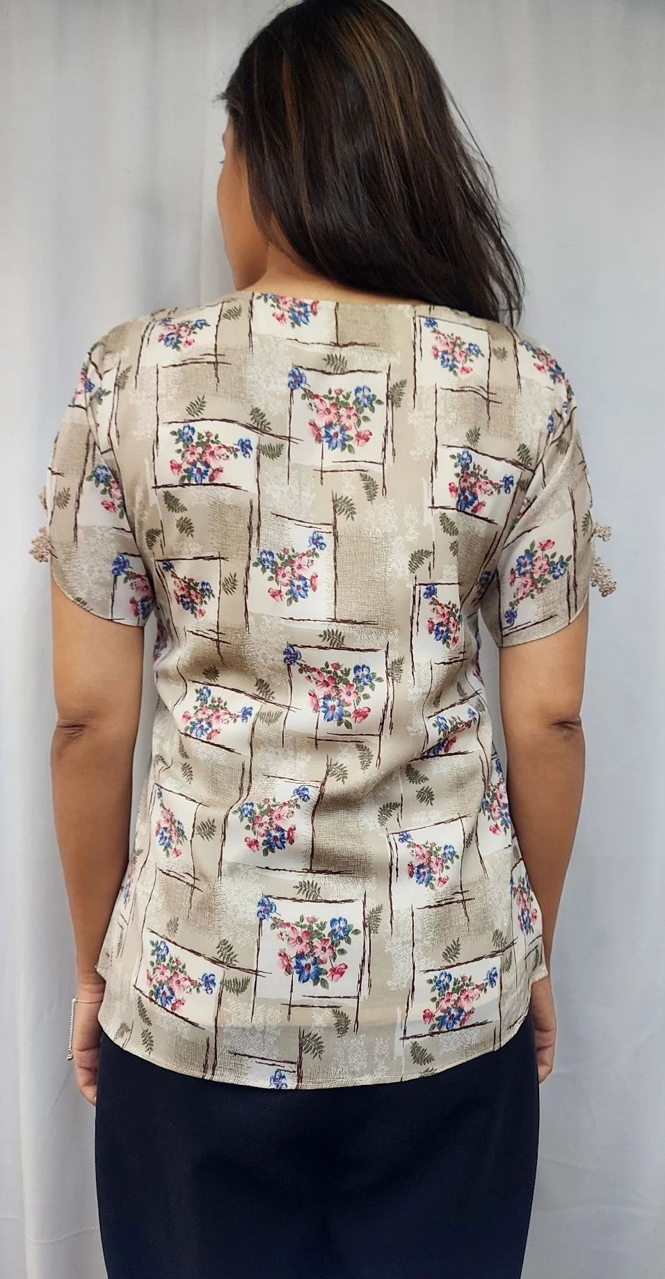 A woman viewed from behind wearing a beige short-sleeve blouse with a floral and geometric print, paired with dark navy pants, standing against a white curtain background.