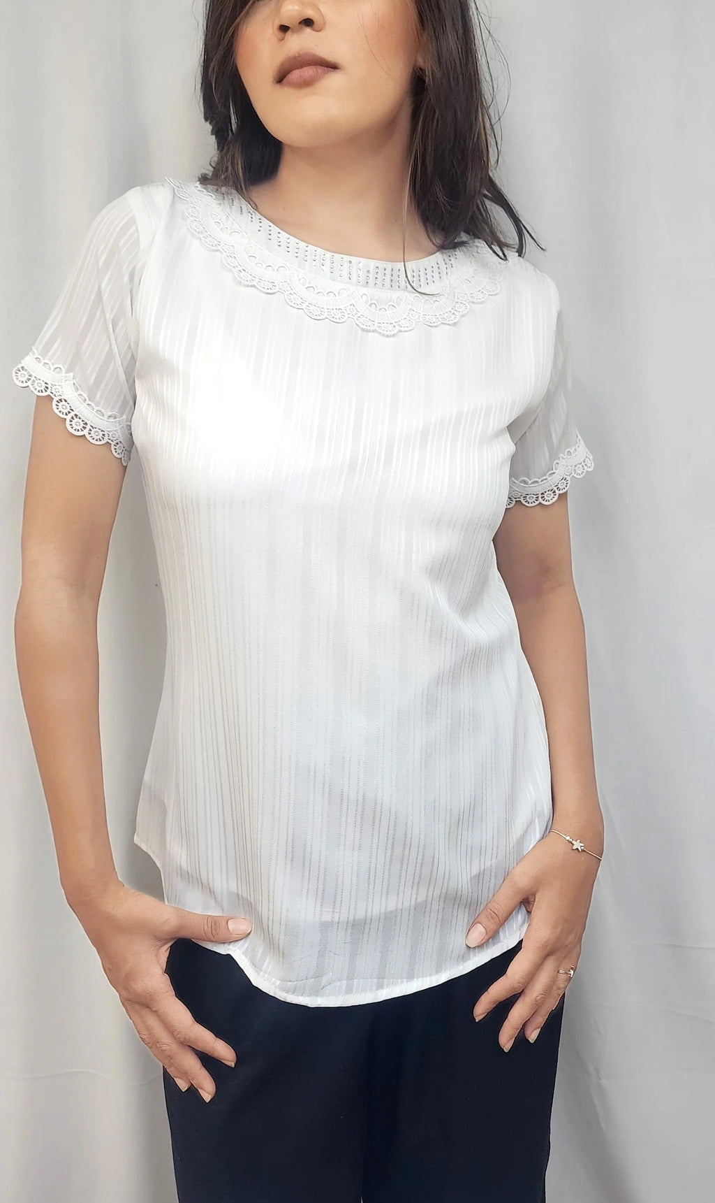 A woman modeling a white short-sleeve blouse with delicate lace trim at the collar and cuffs, paired with dark pants, standing against a light gray fabric backdrop.
