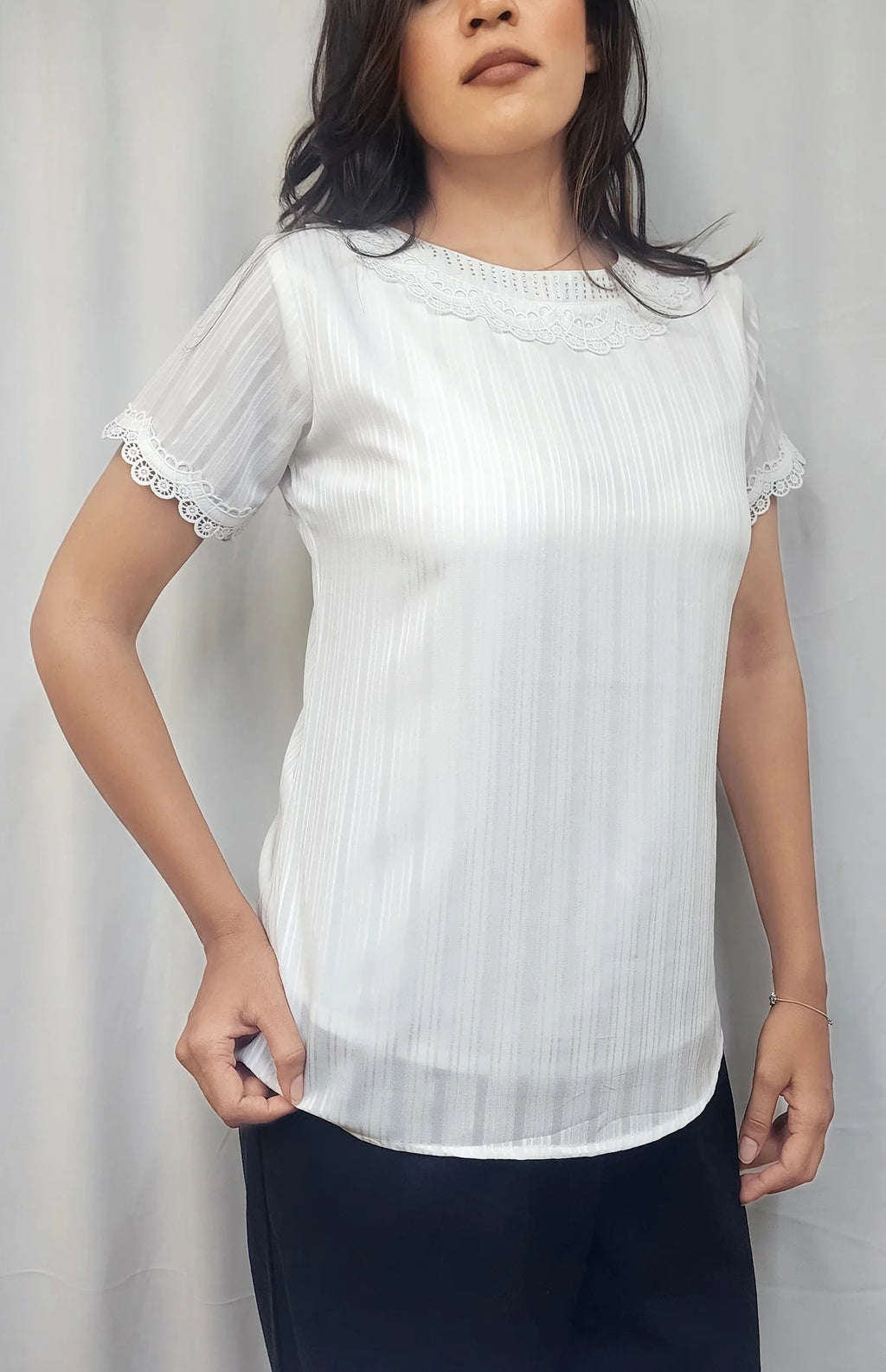 A woman modeling a white short-sleeve blouse with subtle vertical pleats and delicate lace trim at the sleeves, paired with dark pants, against a light gray curtain backdrop.