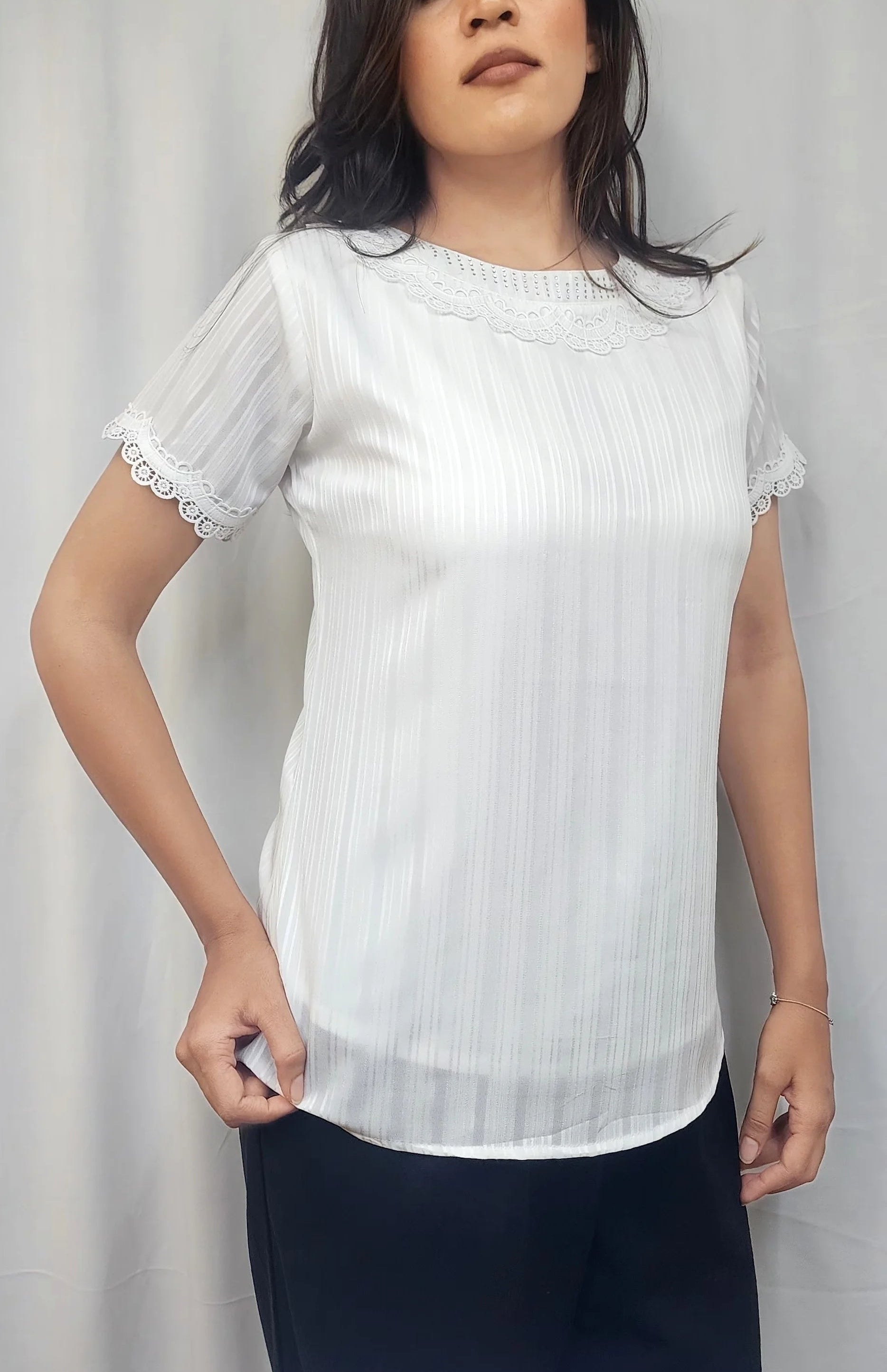 A woman modeling a white short-sleeve blouse with subtle vertical pleats and delicate lace trim at the sleeves, paired with dark pants, against a light gray curtain backdrop.