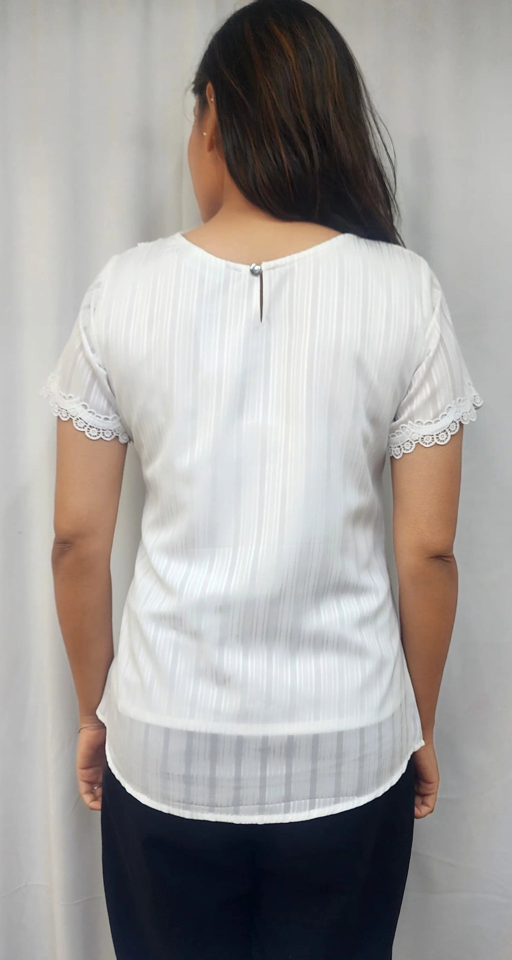 A woman viewed from the back wearing a white short-sleeve blouse with subtle vertical pinstripes, lace trim at the sleeve cuffs, and a small back zipper, paired with dark pants against a light gray curtain backdrop.