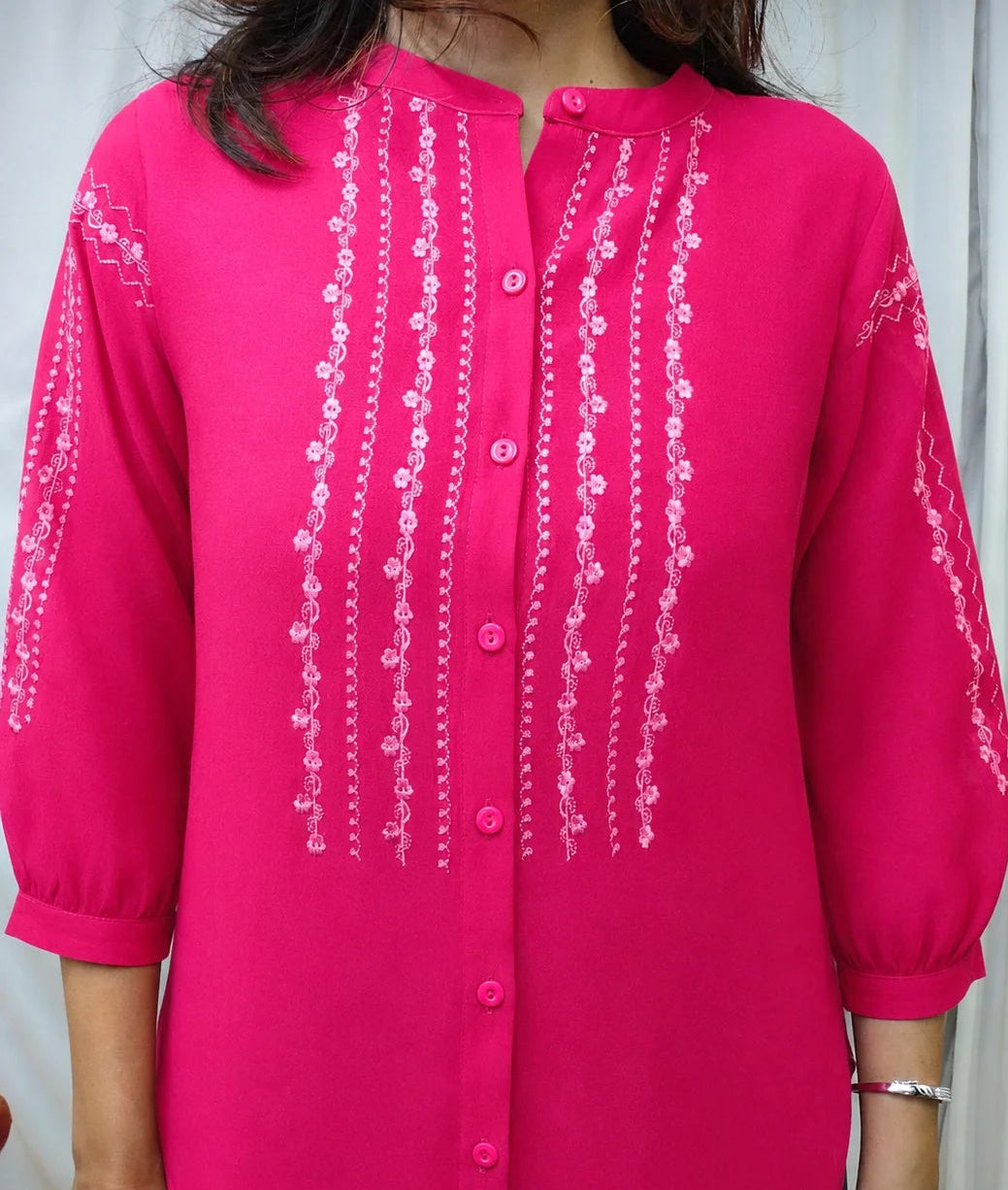 A woman wearing a bright pink button-up blouse with white embroidered vertical patterns on the front and sleeves, shown against a light curtain background.