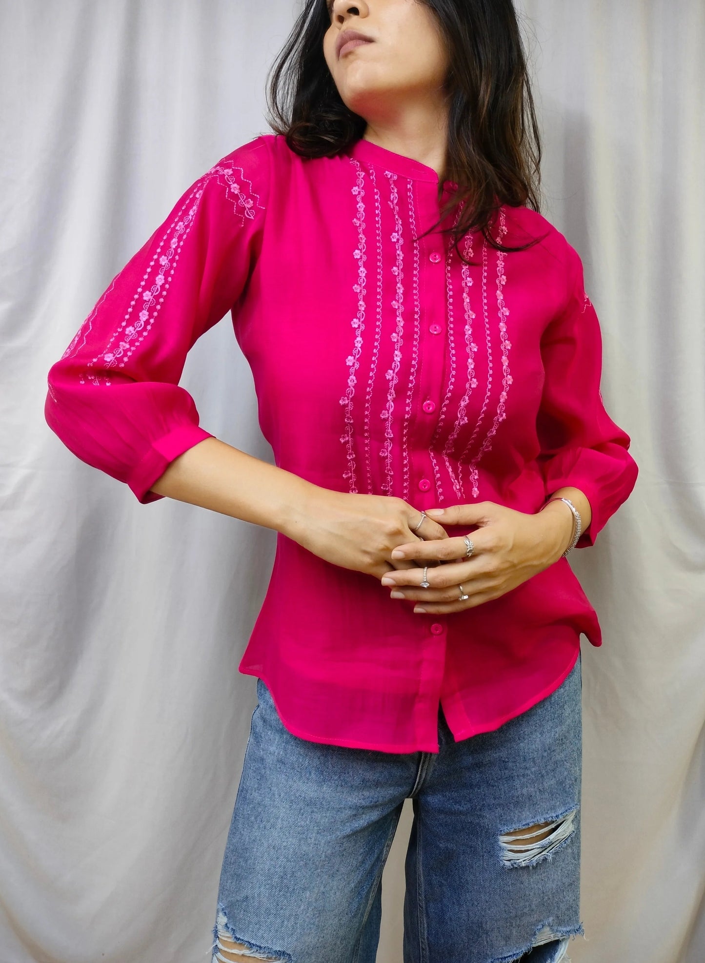 A woman wearing a bright fuchsia pink embroidered long-sleeve blouse and distressed blue jeans, standing in front of a draped white fabric backdrop.