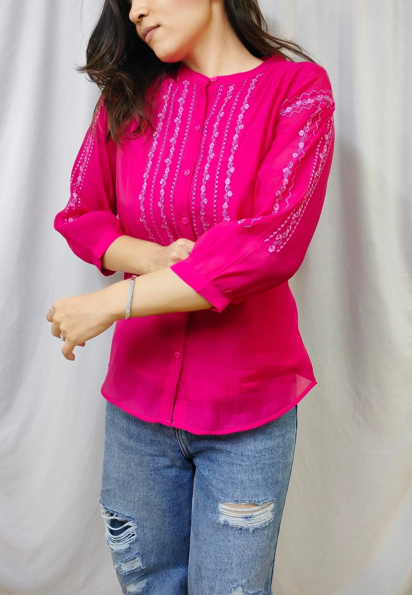 A woman wearing a bright pink blouse with vertical silver mirror embroidery and three-quarter sleeves, paired with distressed blue jeans, standing in front of a draped white fabric backdrop.