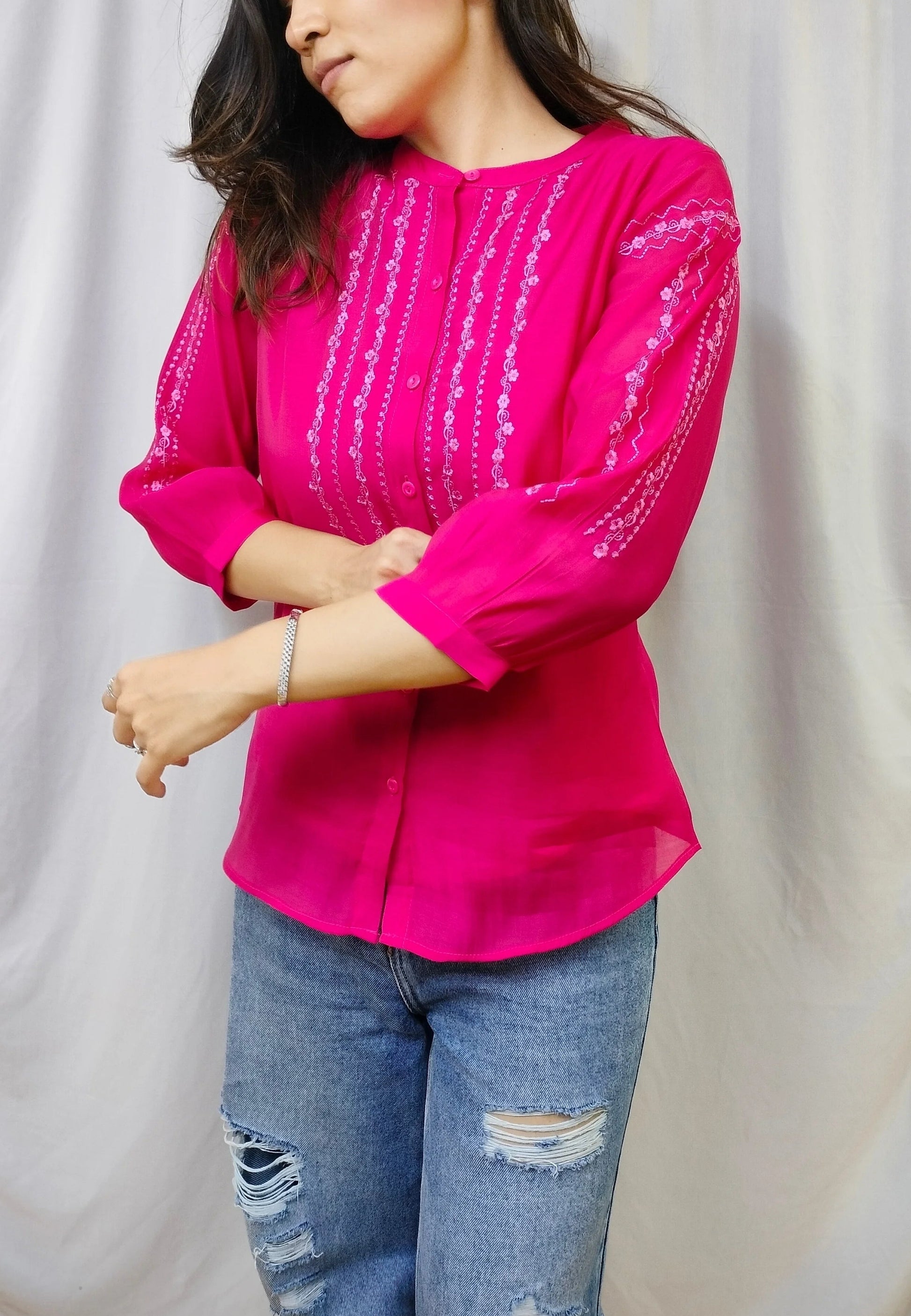 A woman wearing a bright pink blouse with vertical silver mirror embroidery and three-quarter sleeves, paired with distressed blue jeans, standing in front of a draped white fabric backdrop.