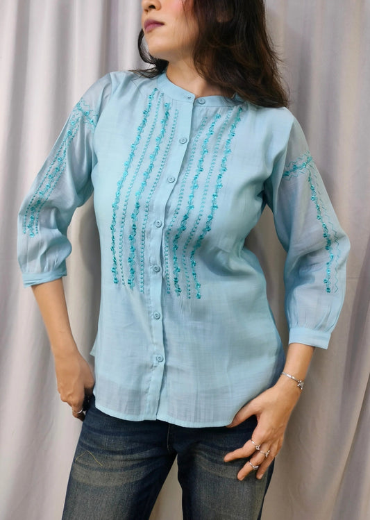 A woman wearing a light blue button-up blouse with turquoise embroidered vertical stripes on the chest and sleeves, paired with dark blue jeans, standing in front of a draped light gray fabric backdrop.