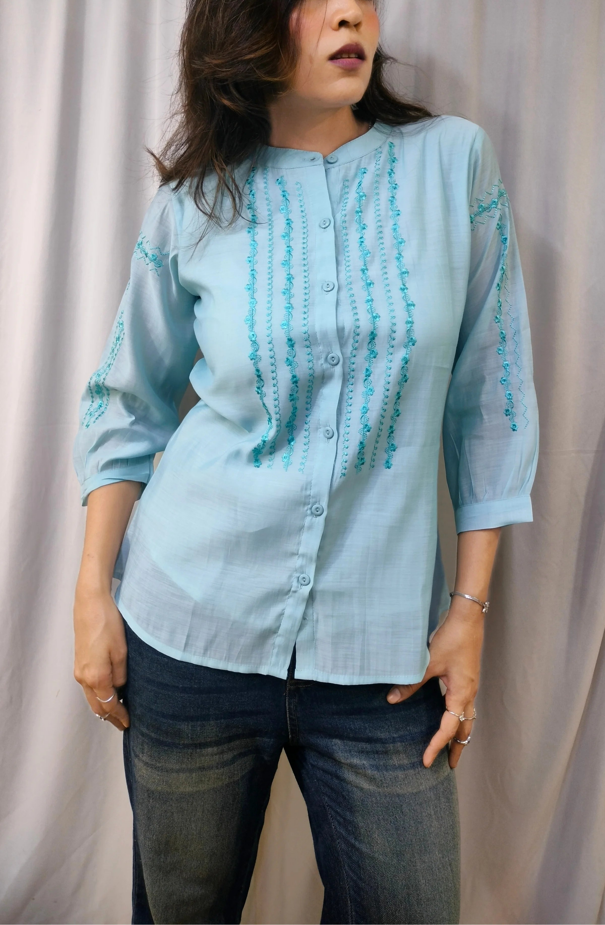 A woman modeling a light blue button-up shirt with turquoise embroidery on the front and sleeves, paired with dark blue jeans, standing against a draped beige fabric backdrop.
