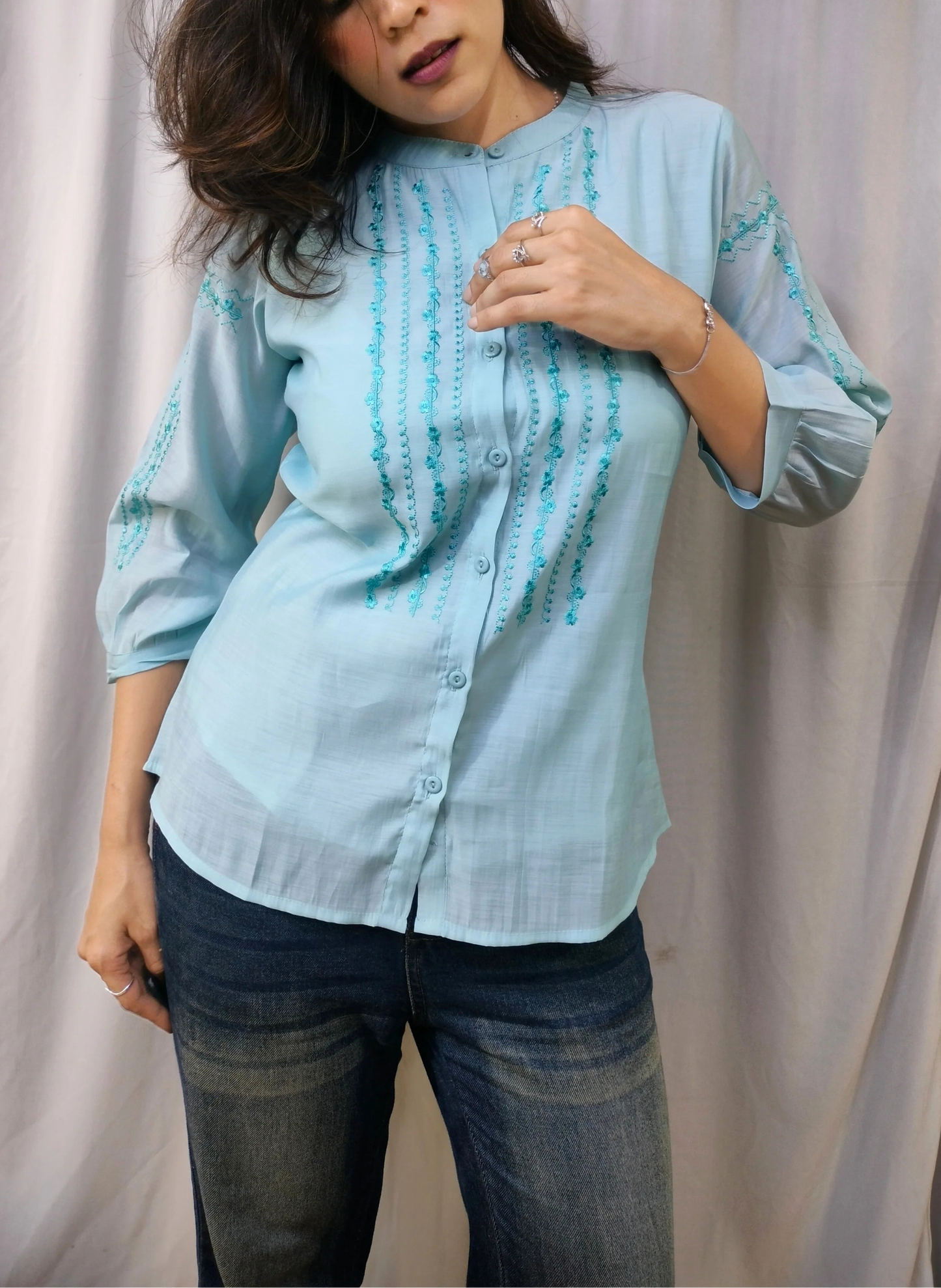 A woman wearing a light blue button-up shirt with vertical teal embroidery and dark blue jeans, posing in front of a draped beige fabric backdrop.