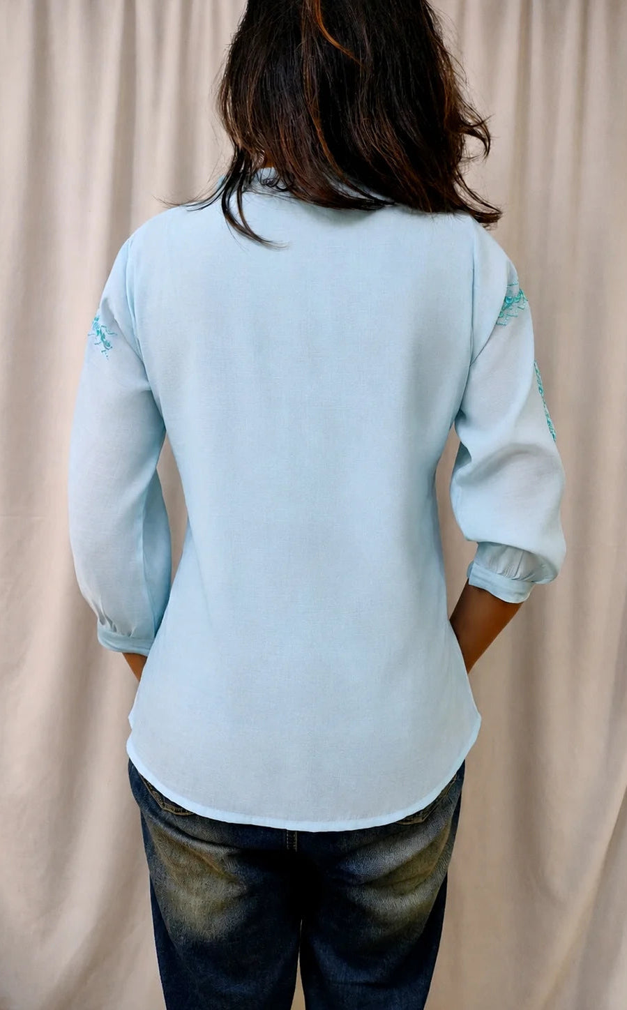 A woman viewed from behind wearing a light blue long-sleeve blouse with subtle teal embroidery on the upper sleeves, paired with dark blue jeans, set against a draped beige fabric backdrop.