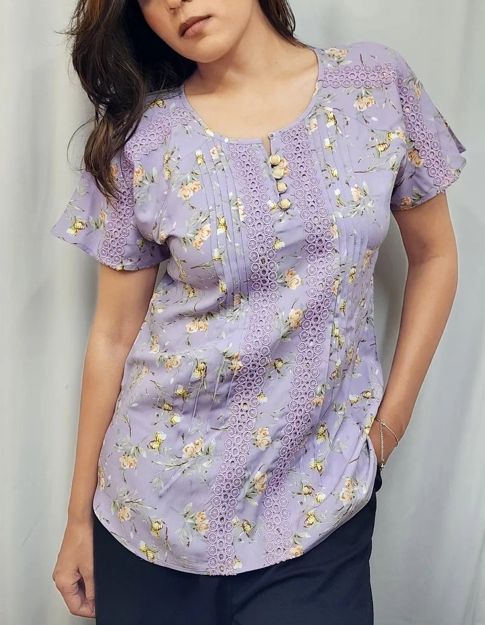 A woman wearing a lavender short-sleeve blouse with a yellow and white floral print, paired with dark pants, standing against a light gray curtain backdrop.