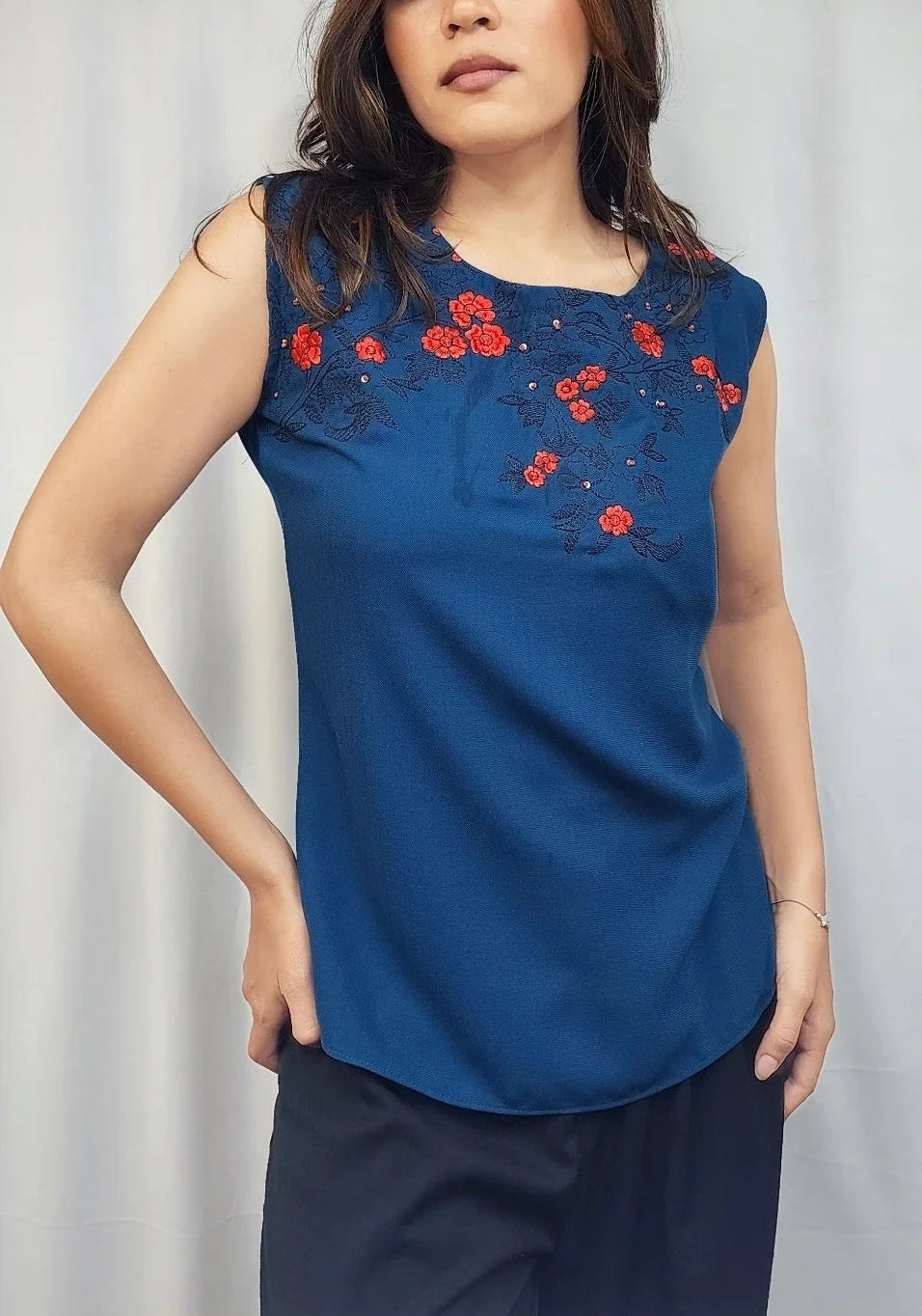 A woman modeling a sleeveless royal blue top with red floral embroidery on the chest, paired with dark navy pants, set against a light gray curtain backdrop.