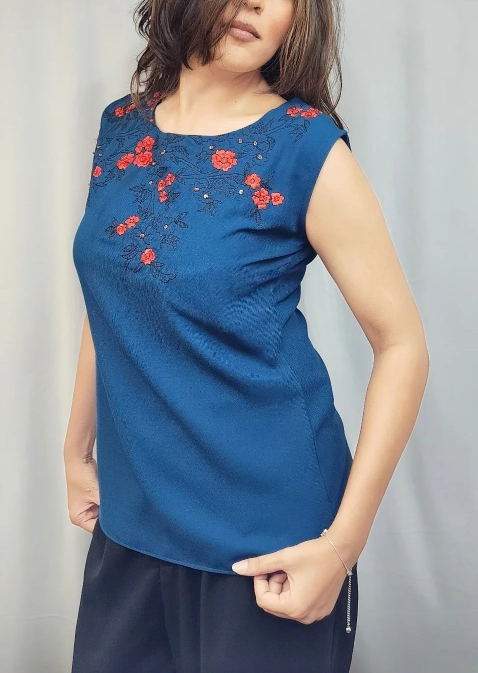 A woman modeling a sleeveless royal blue top with red floral embroidery on the chest, paired with dark navy bottoms, standing in front of a light gray curtain.
