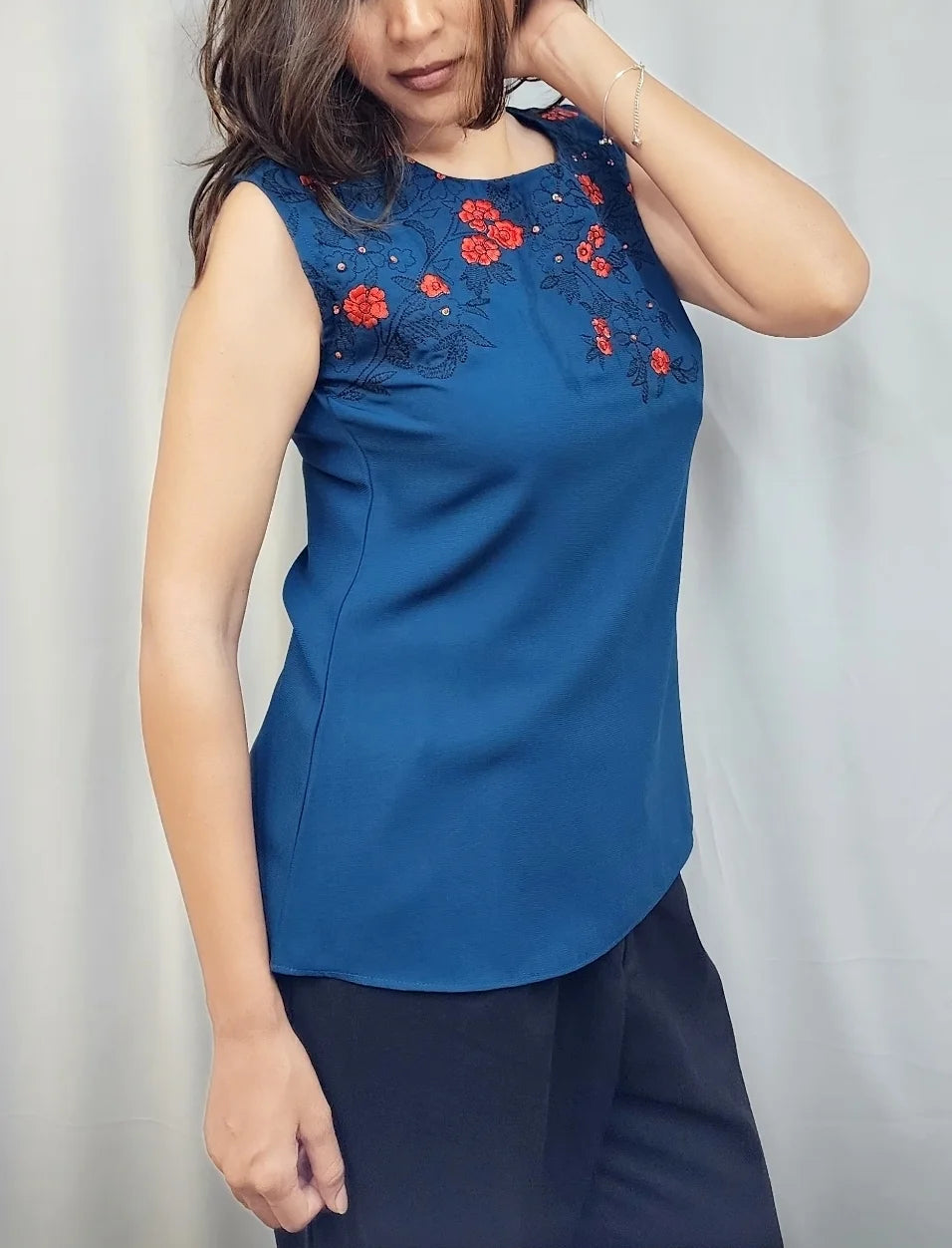 A woman modeling a sleeveless blue top with red floral embroidery on the chest, paired with dark pants, standing in front of a light gray curtain backdrop.