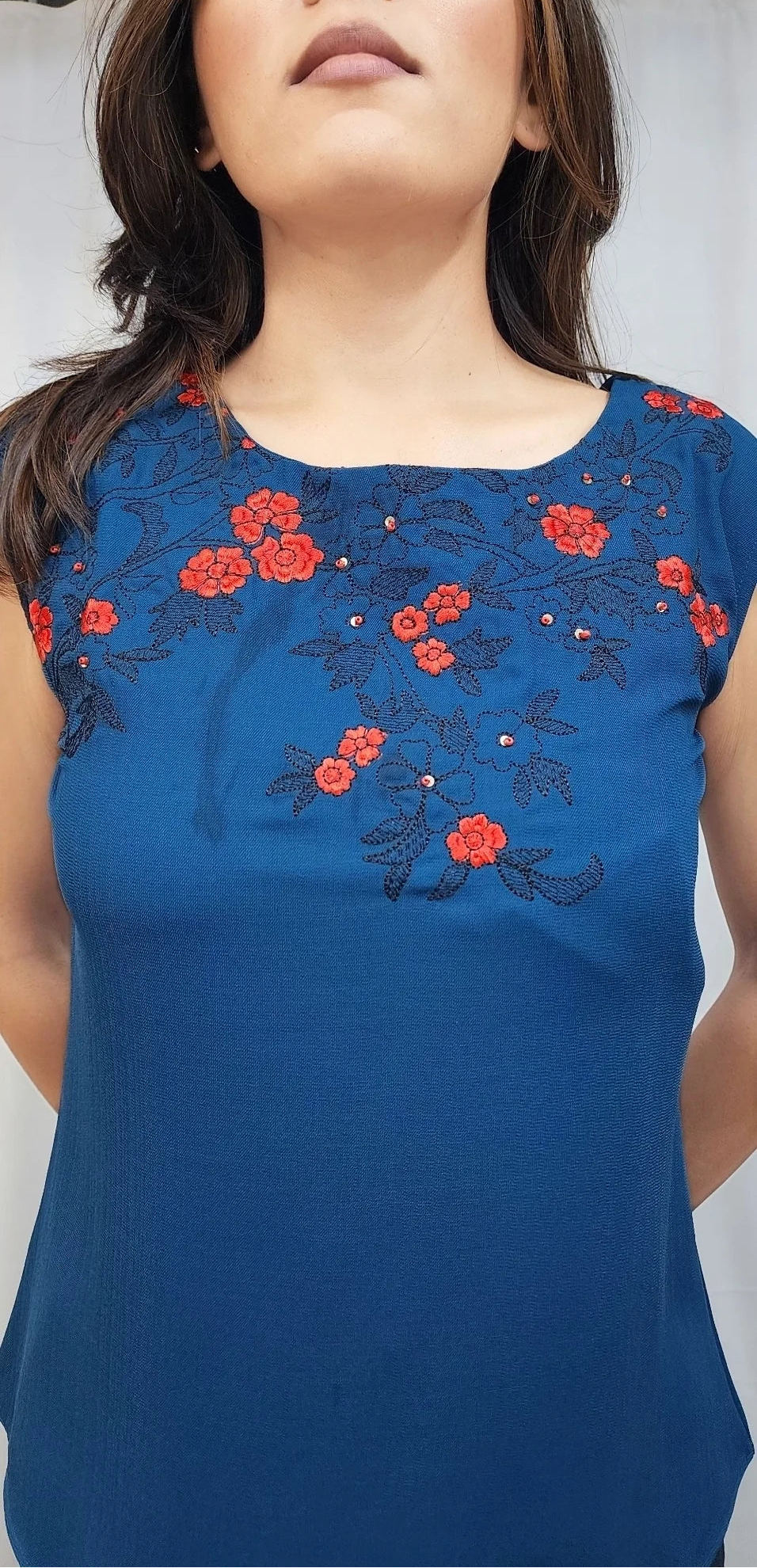 A woman wearing a sleeveless royal blue top with red floral embroidery across the chest, shown from the shoulders to the waist against a light gray background.