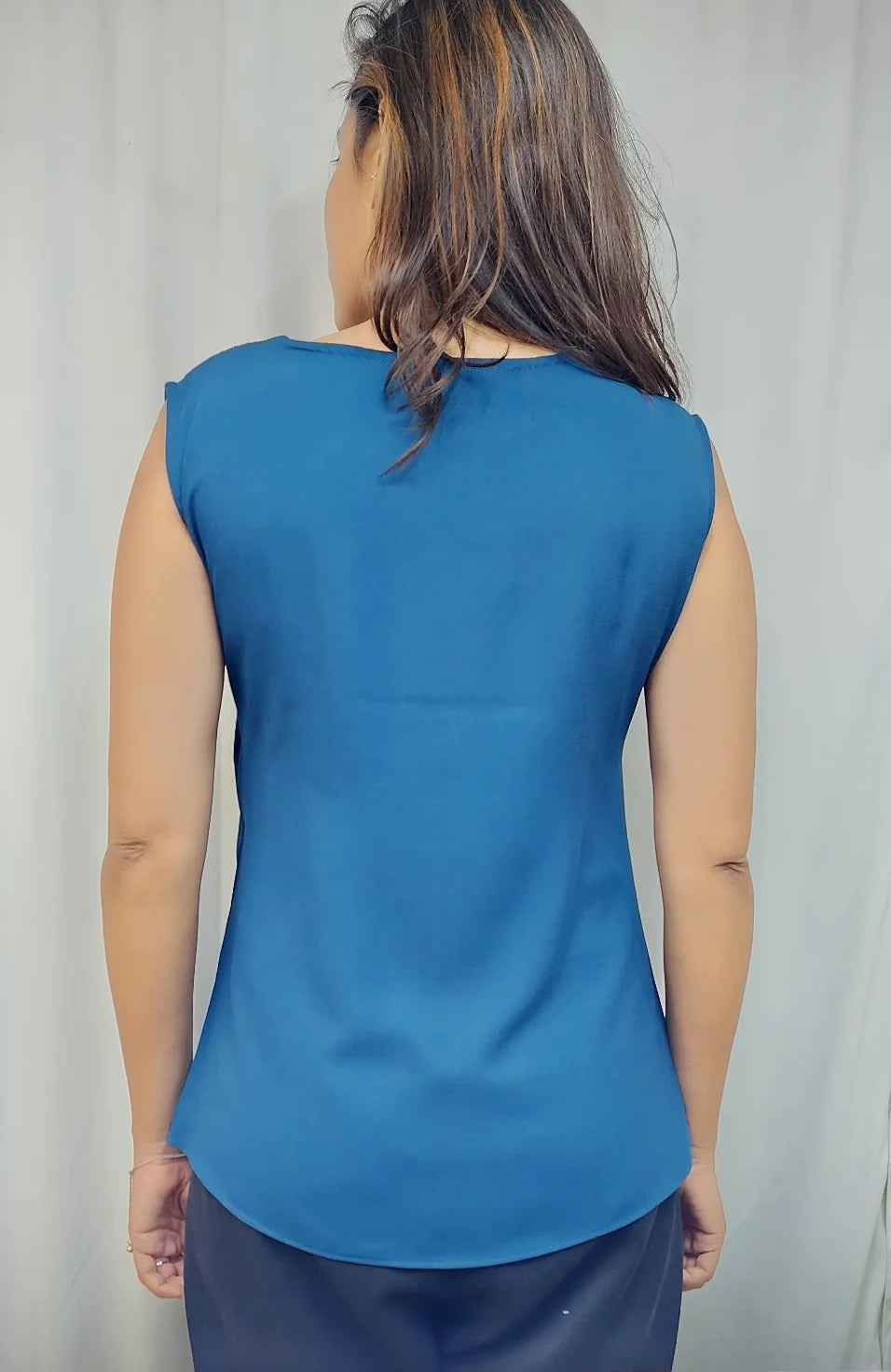 A woman viewed from behind wearing a sleeveless blue top and dark bottoms, standing against a light curtain backdrop.