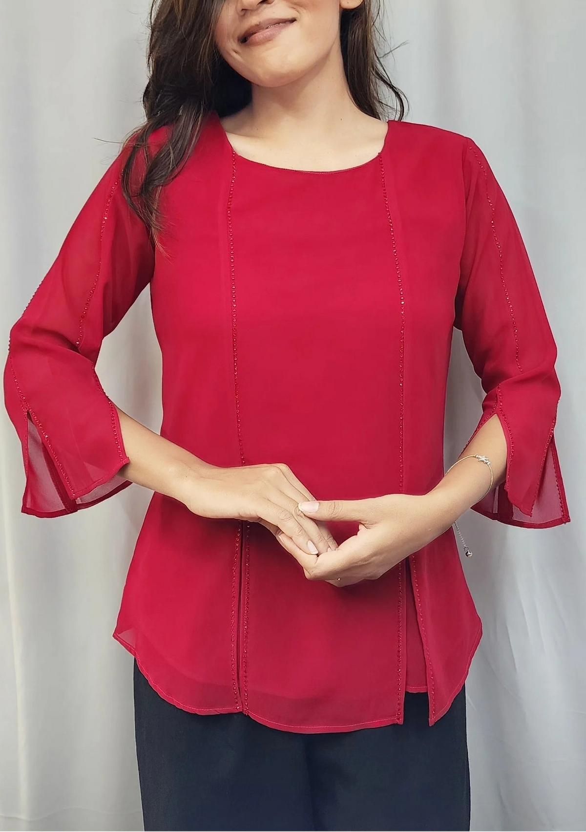 A woman modeling a bright red three-quarter sleeve blouse with flared bell sleeves and a round neckline, paired with dark pants, standing in front of a white curtain.