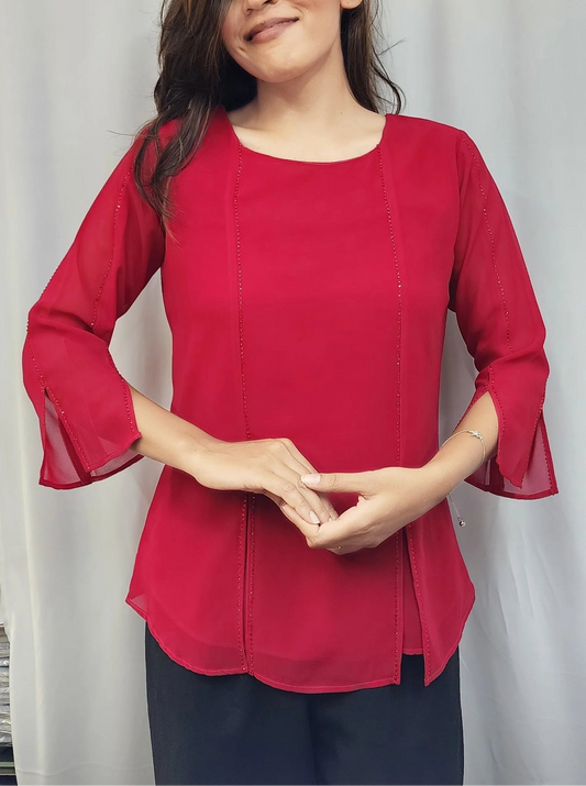 A woman modeling a bright red three-quarter sleeve blouse with flared bell sleeves and a round neckline, paired with dark pants, standing in front of a white curtain.