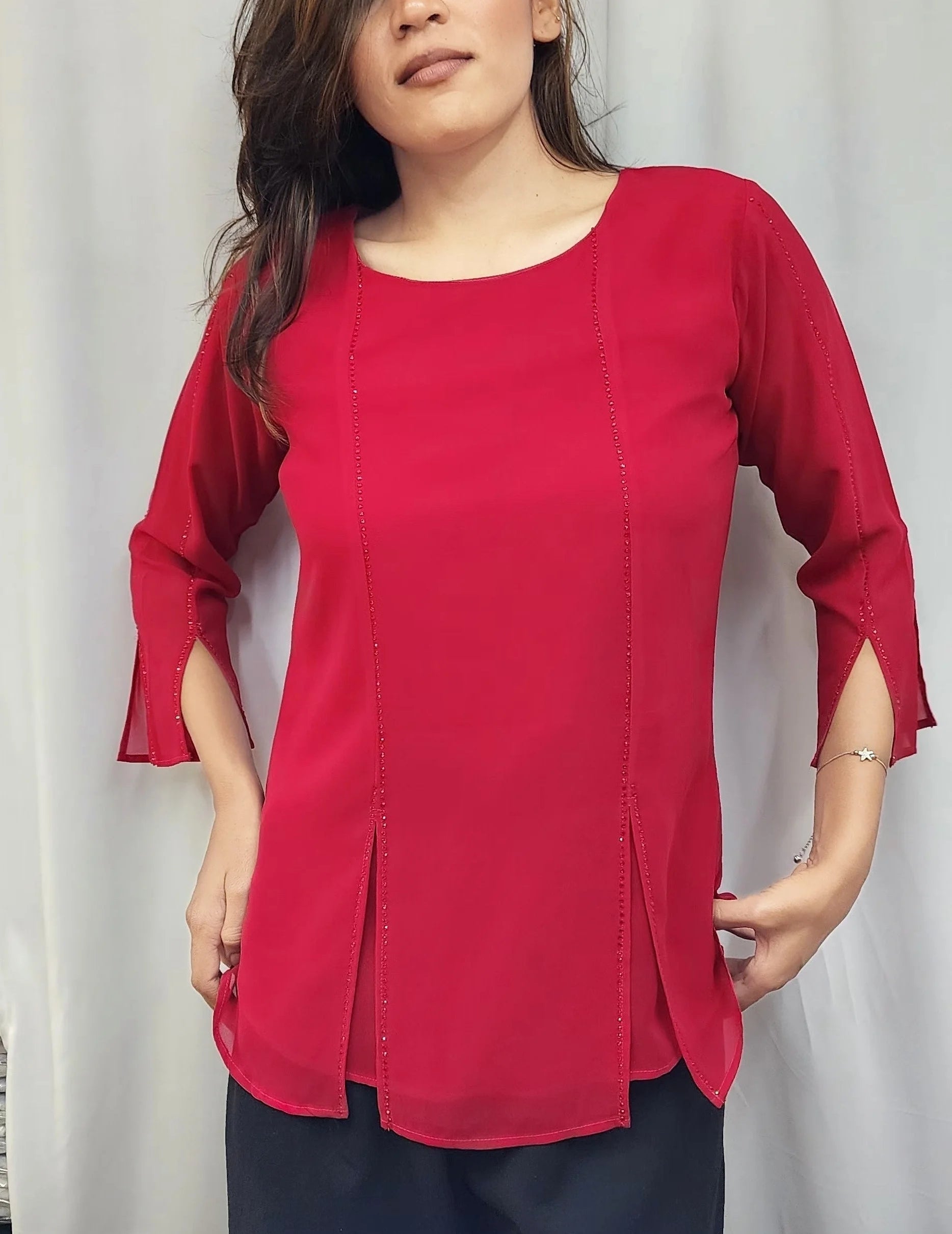 A woman modeling a bright red three-quarter sleeve top with a round neckline and side slits, paired with dark pants, standing in front of a white curtain backdrop.