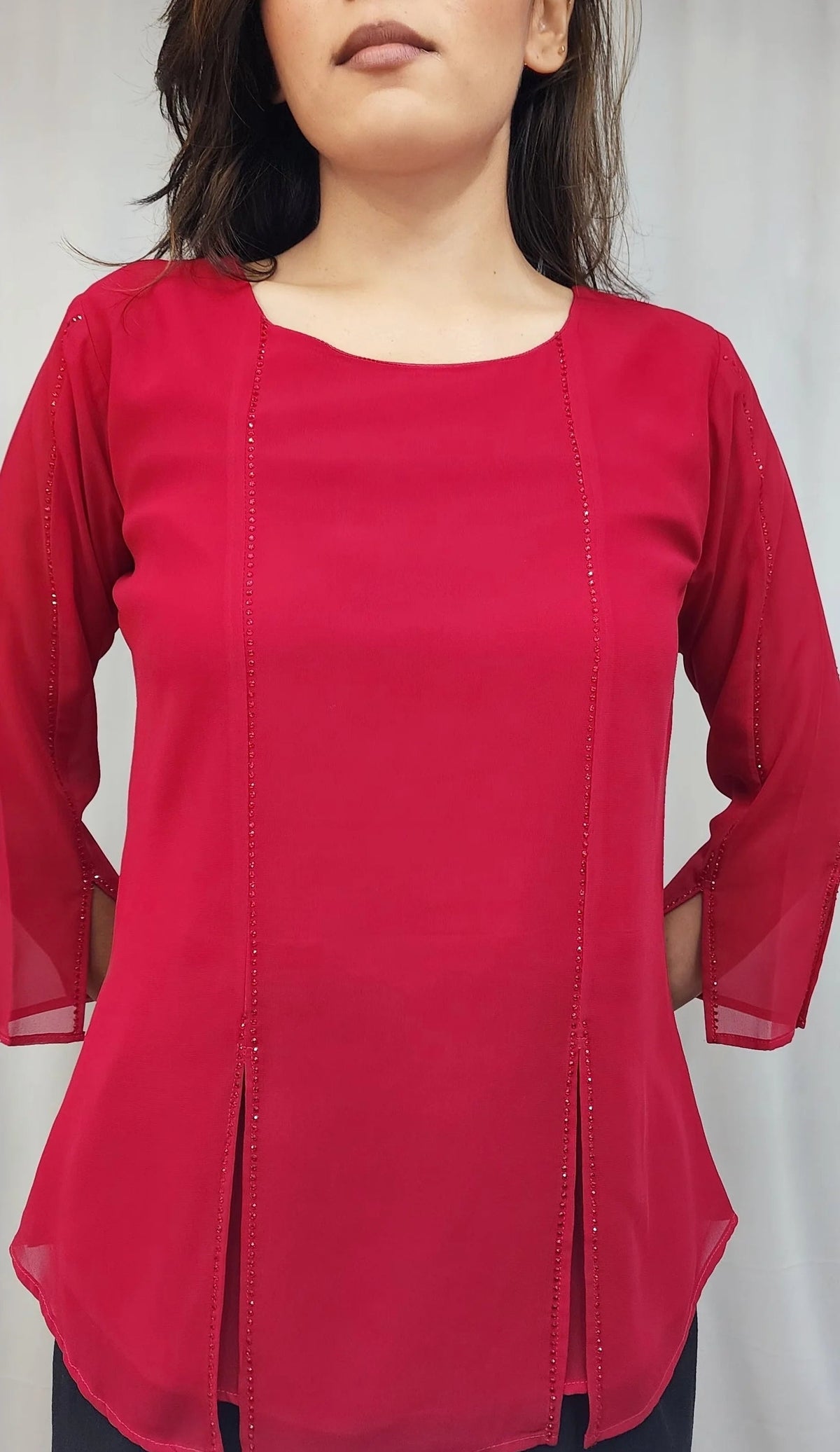 A woman wearing a bright red three-quarter sleeve top with vertical seam detailing, photographed against a light gray curtain background.
