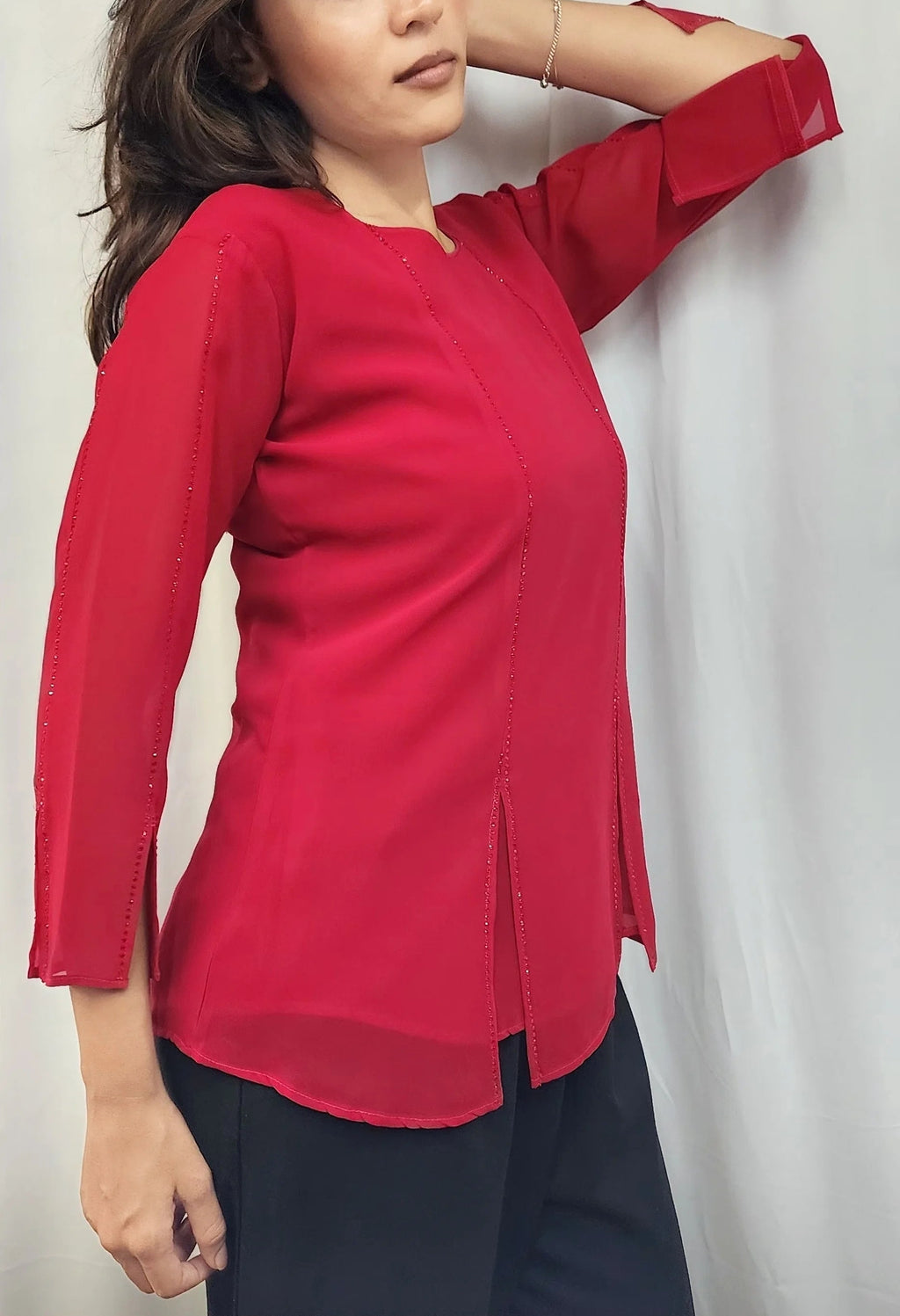 A woman wearing a bright red three-quarter sleeve top with a split hem, paired with dark pants, posing against a light gray curtain backdrop.