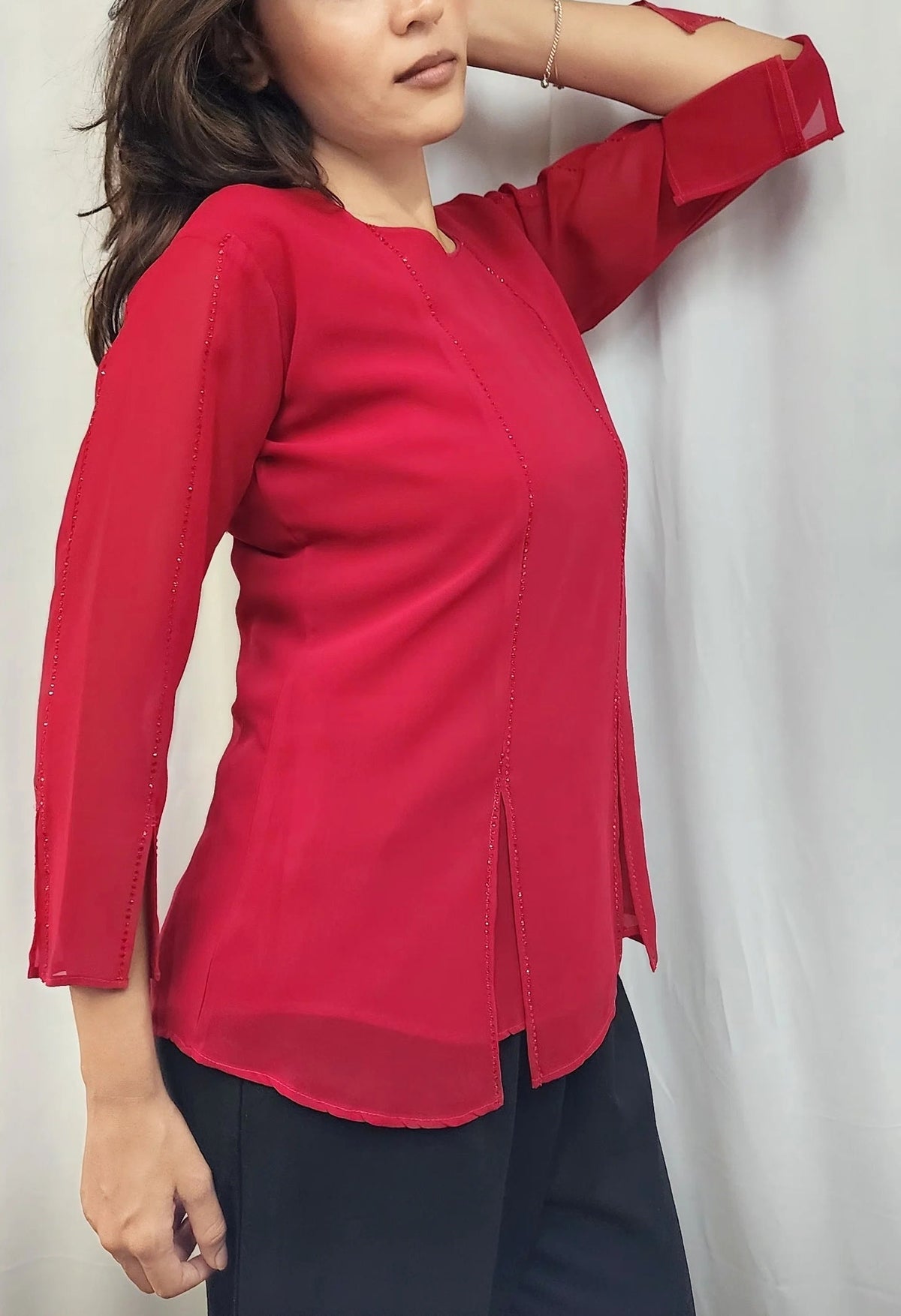A woman wearing a bright red three-quarter sleeve top with a split hem, paired with dark pants, posing against a light gray curtain backdrop.