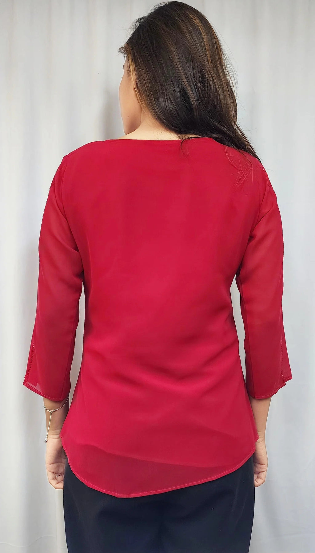 A woman viewed from the back wearing a bright red three-quarter sleeve top and dark navy pants, standing in front of a white curtain.