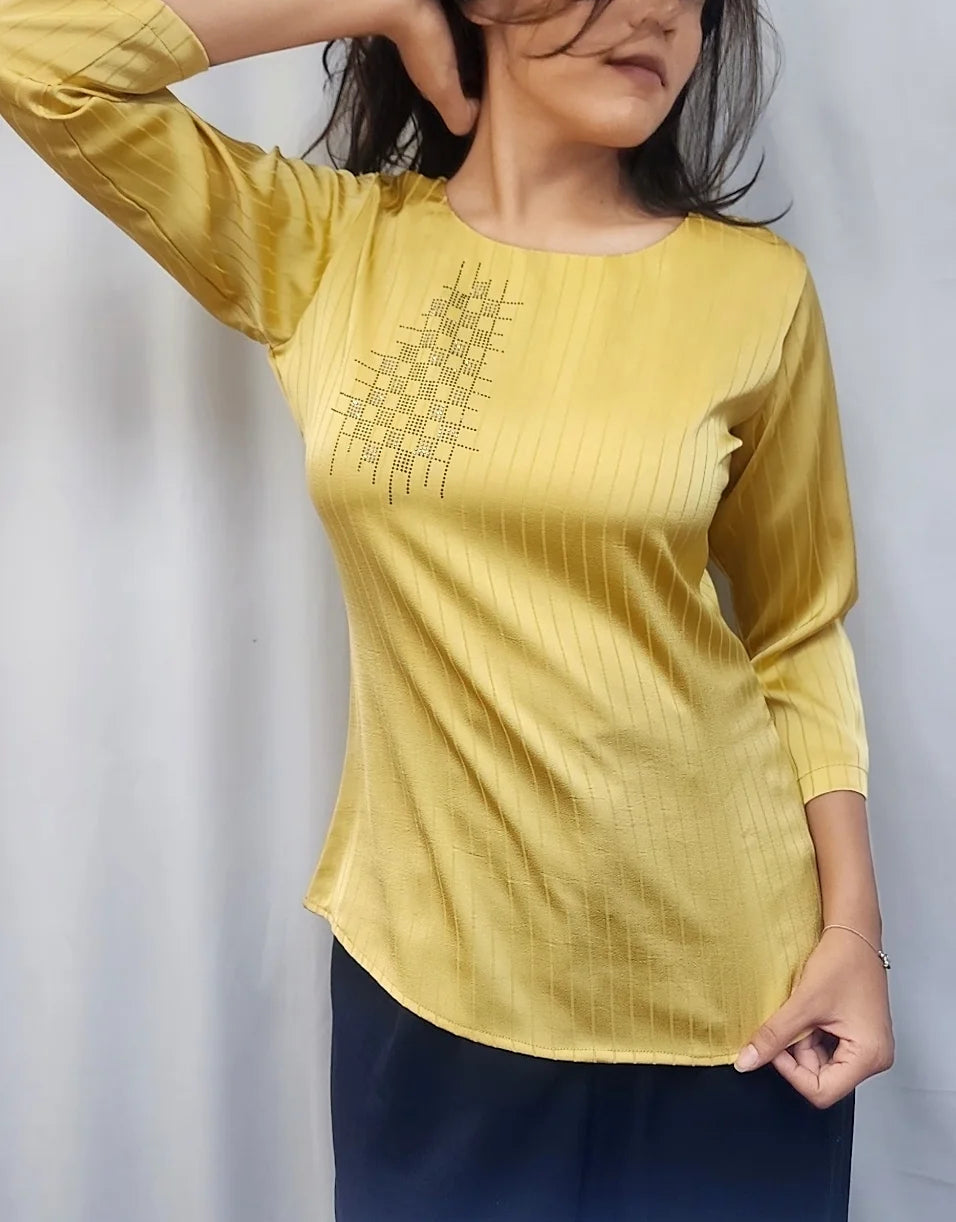 A woman modeling a yellow long-sleeve top with a geometric embroidered pattern on the chest, paired with dark navy pants, set against a white curtain backdrop.