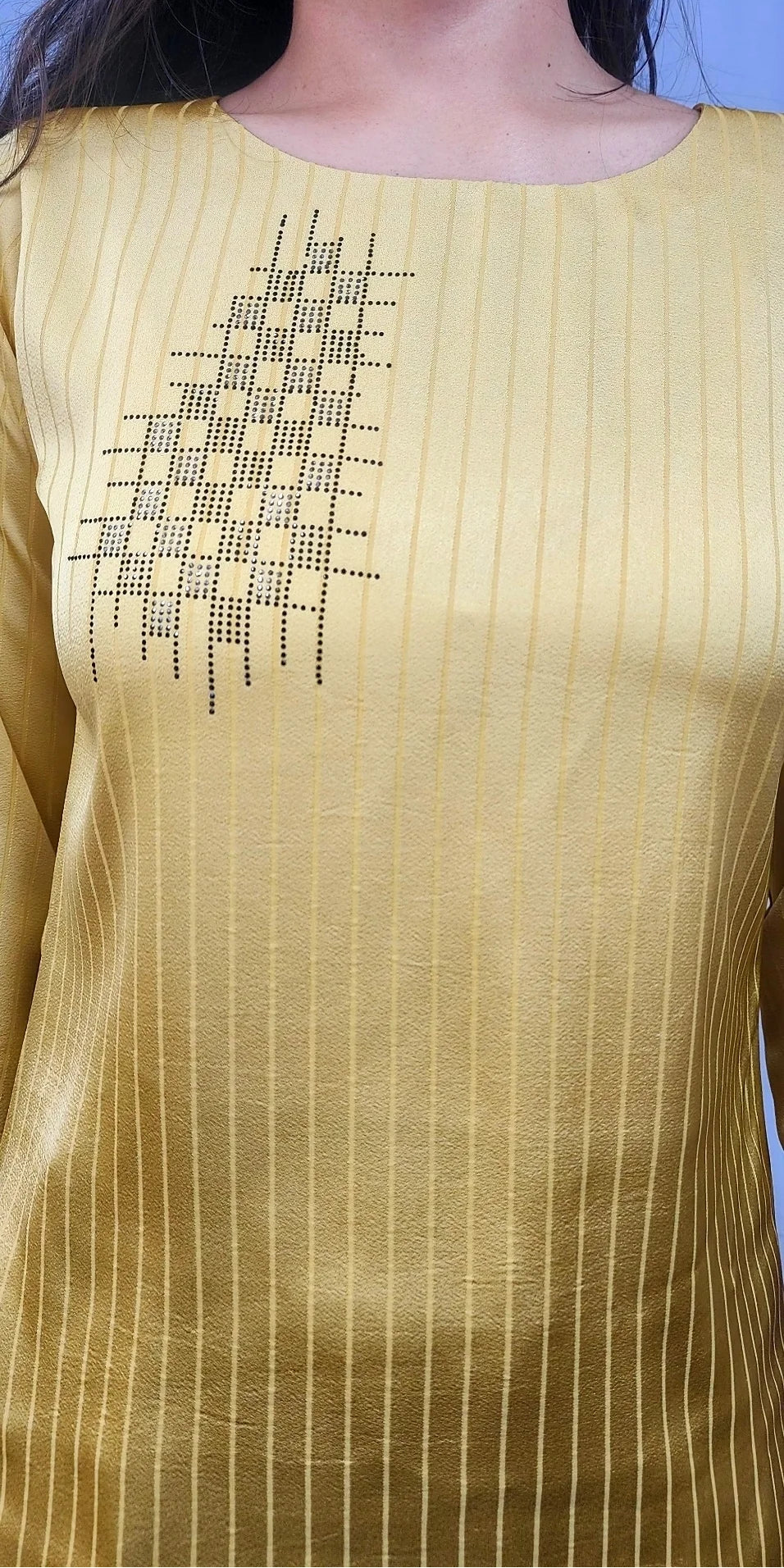 A close-up of a person wearing a pale yellow top with subtle vertical pinstripes and a black geometric embroidered pattern on the chest.