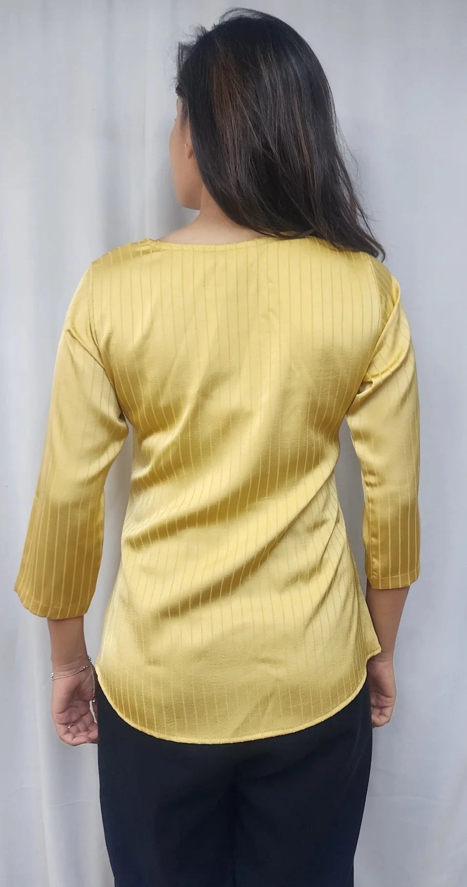A woman viewed from the back wearing a yellow ribbed three-quarter sleeve top and dark pants, standing against a light gray curtain backdrop.