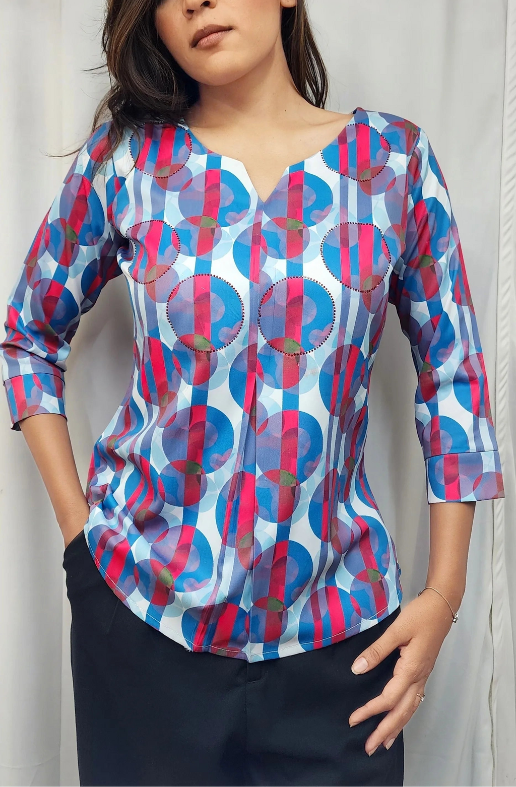A woman modeling a three-quarter sleeve top with a vibrant blue, pink, and red circular geometric pattern on a white base, paired with black trousers, set against a light gray curtain background.