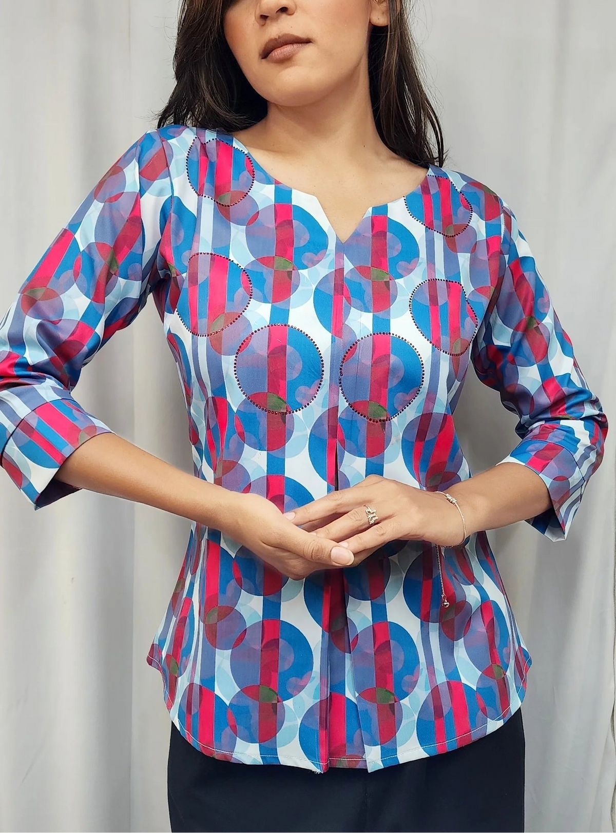 A woman modeling a long-sleeve V-neck blouse with a blue, pink, and white geometric pattern, paired with dark pants, set against a light gray curtain background.