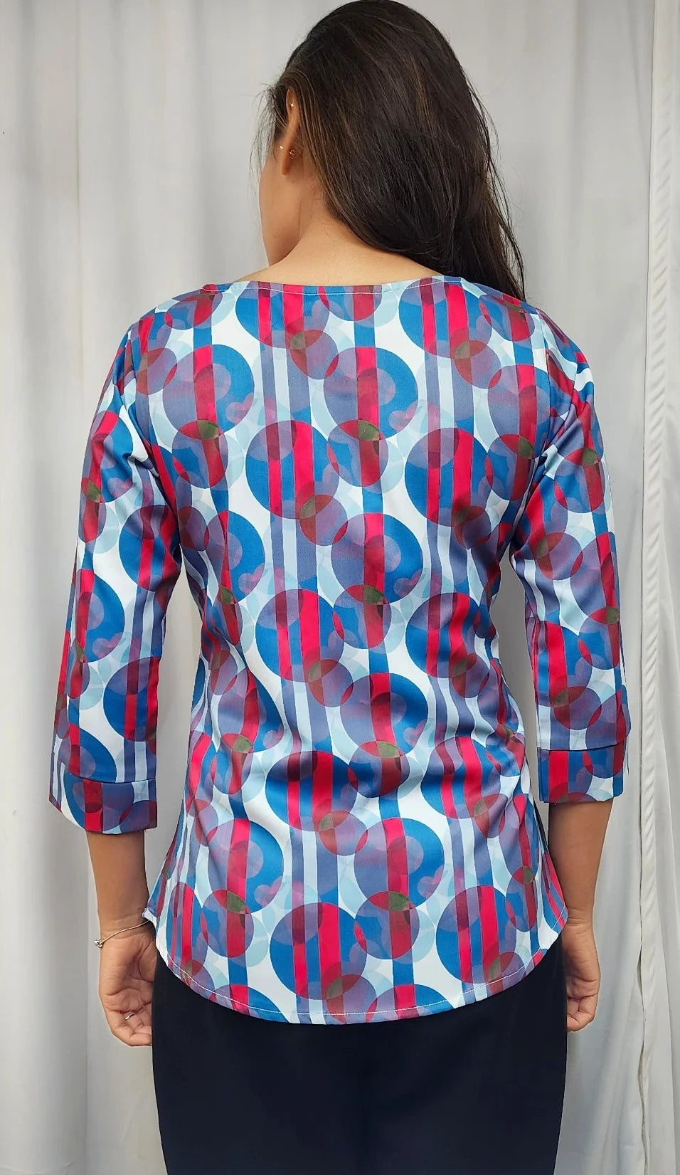 A woman viewed from behind wearing a three-quarter sleeve top with a colorful geometric pattern of blue, red, and pink circles and vertical stripes on a white base, paired with a dark skirt.