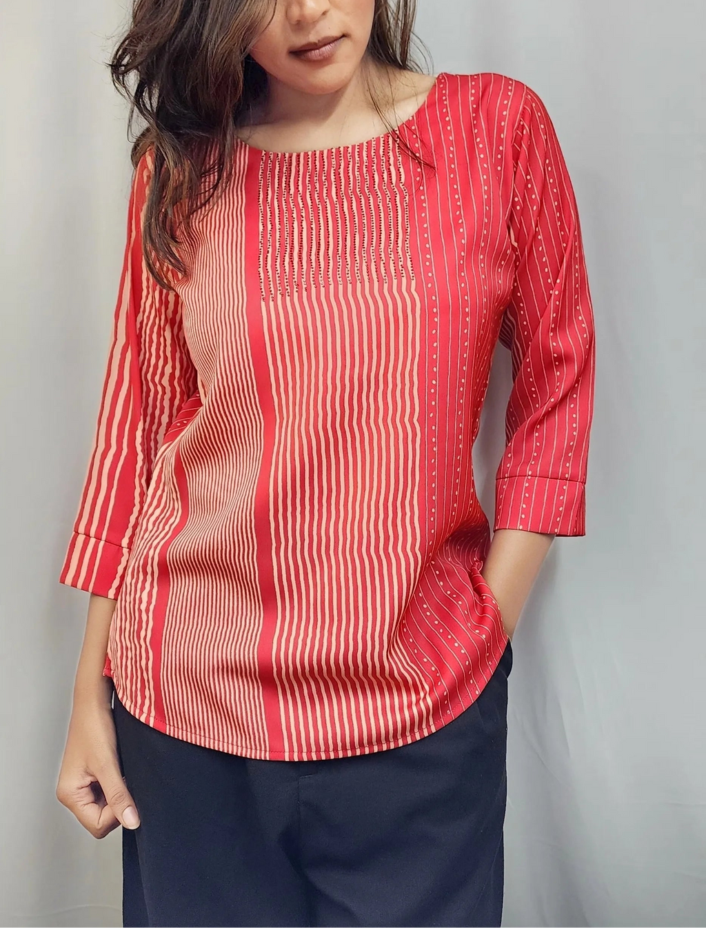 A woman wearing a red and white vertically striped three-quarter sleeve top with a round neckline, paired with dark navy pants, standing against a light gray curtain backdrop.