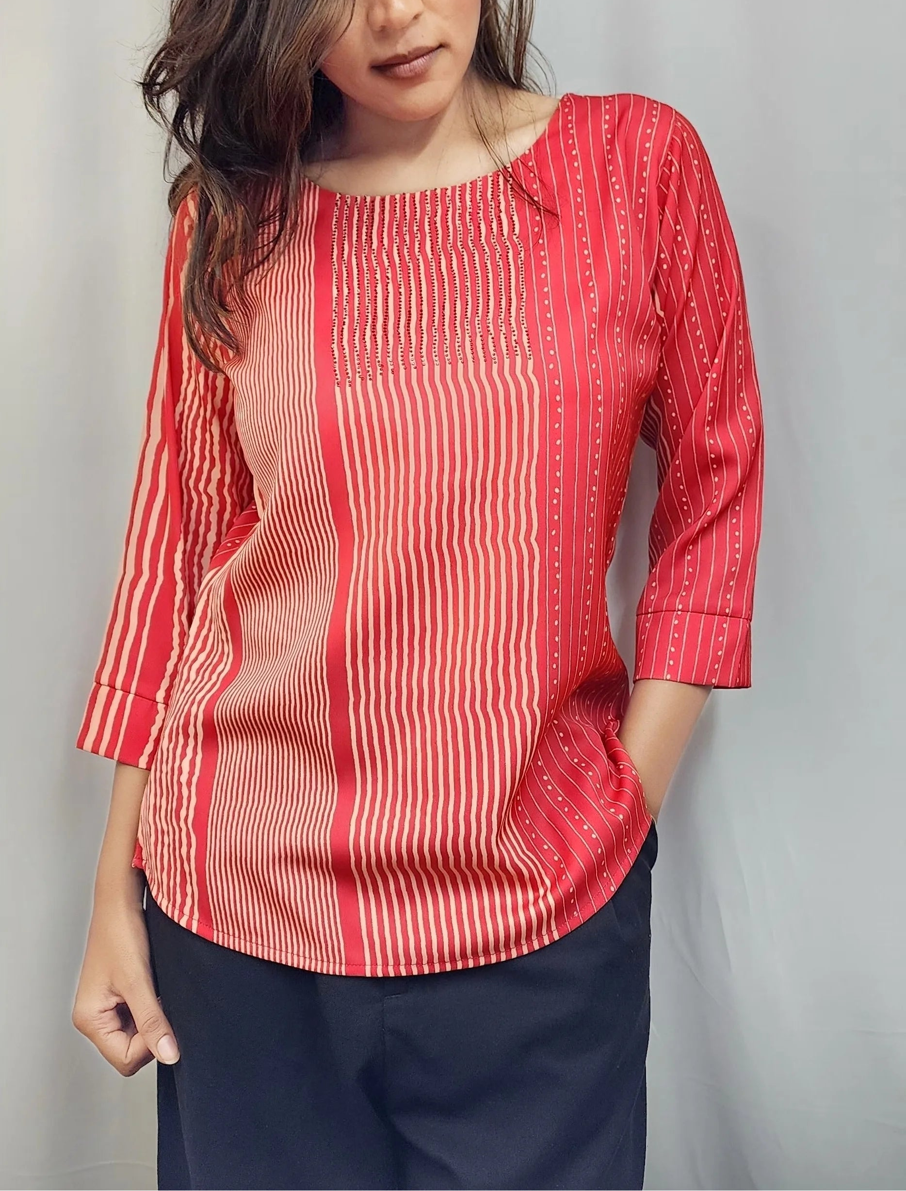 A woman wearing a red and white vertically striped three-quarter sleeve top with a round neckline, paired with dark navy pants, standing against a light gray curtain backdrop.