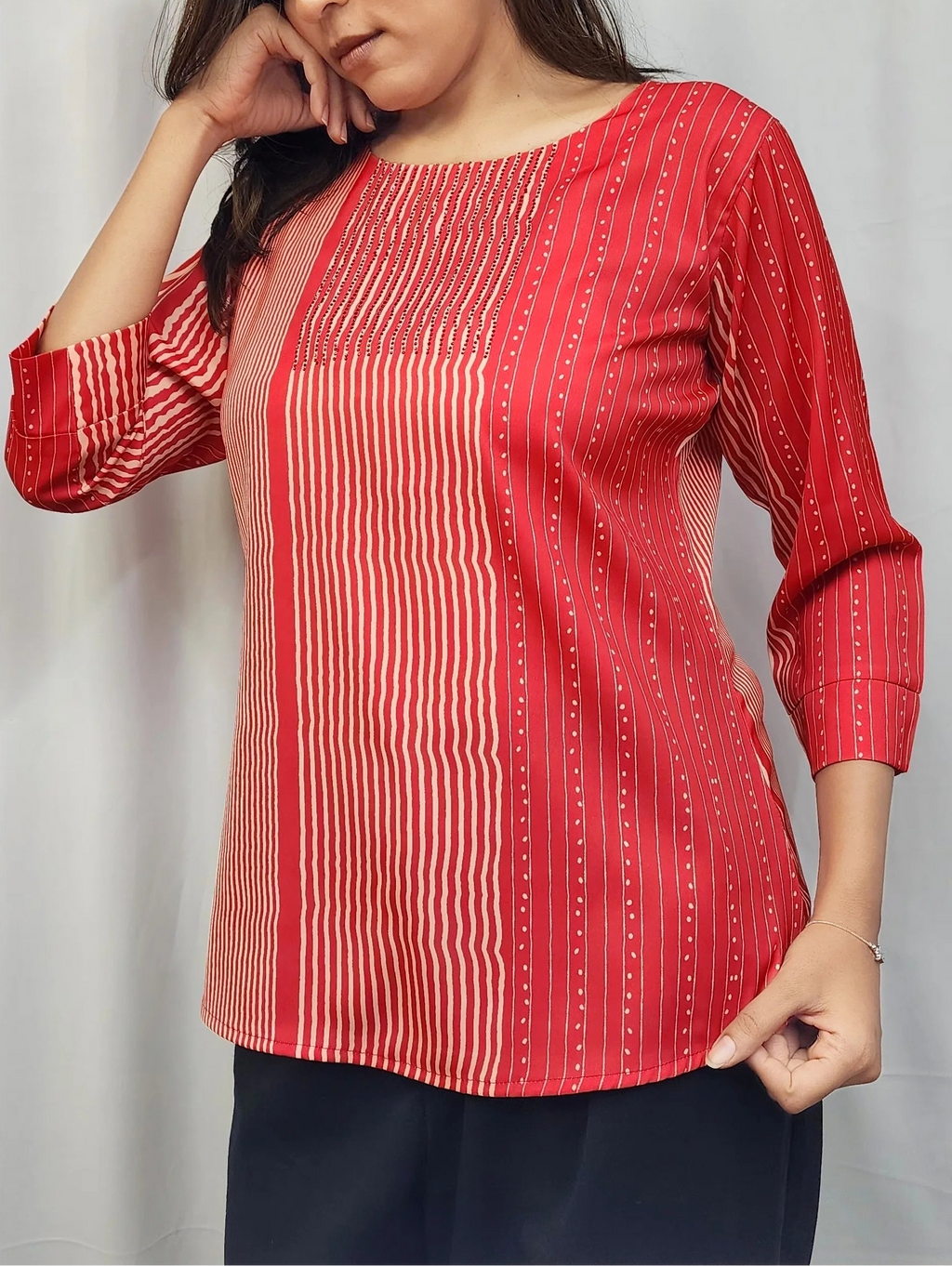 A woman wearing a red three-quarter sleeve top with vertical white stripes and a small white dot pattern, paired with dark navy pants, standing against a light gray curtain background.