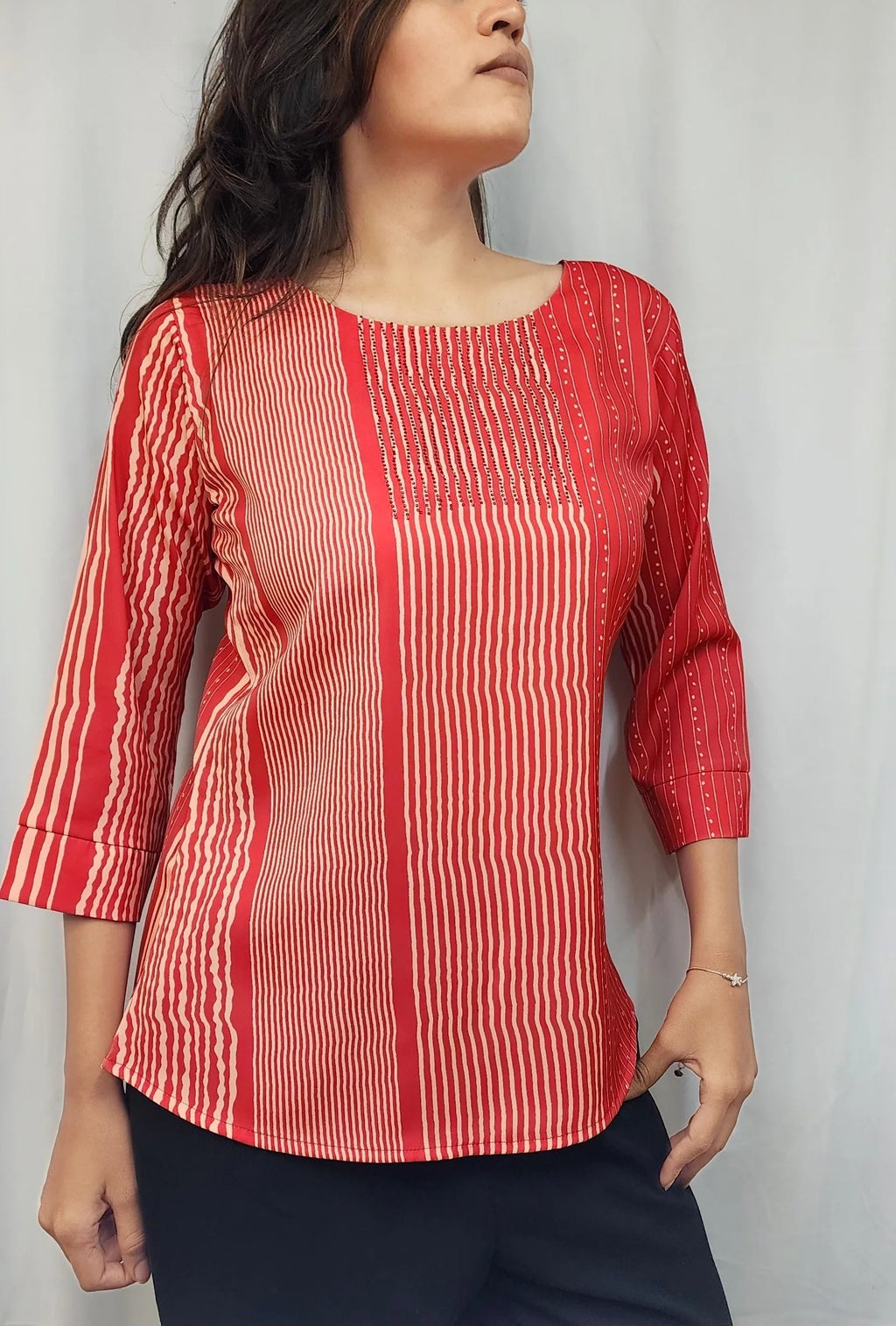 A woman wearing a red and white vertically striped three-quarter sleeve top, paired with black pants, standing against a light gray curtain backdrop.