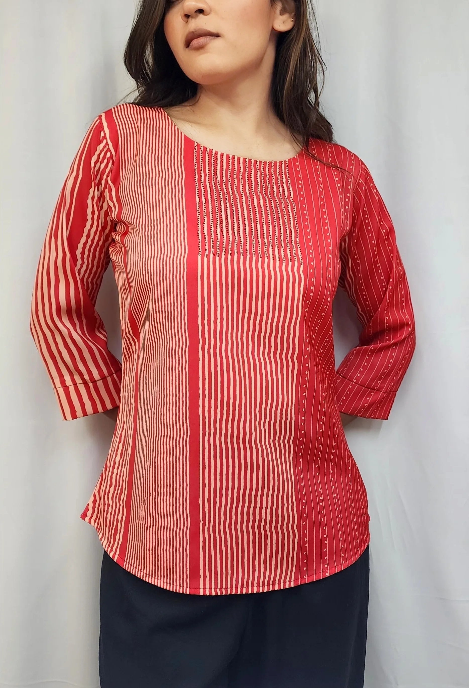 A woman wearing a red and white vertically striped long-sleeve top with a round neckline, paired with dark navy pants, standing against a light gray curtain backdrop.