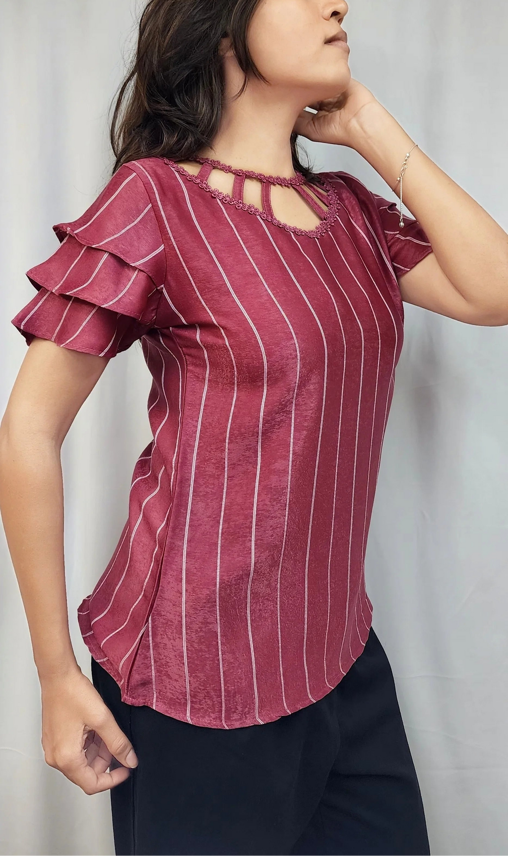 A woman wearing a short-sleeve ruffled blouse with thin vertical white stripes on a dusty rose-red background, paired with dark pants, standing against a light gray curtain backdrop.
