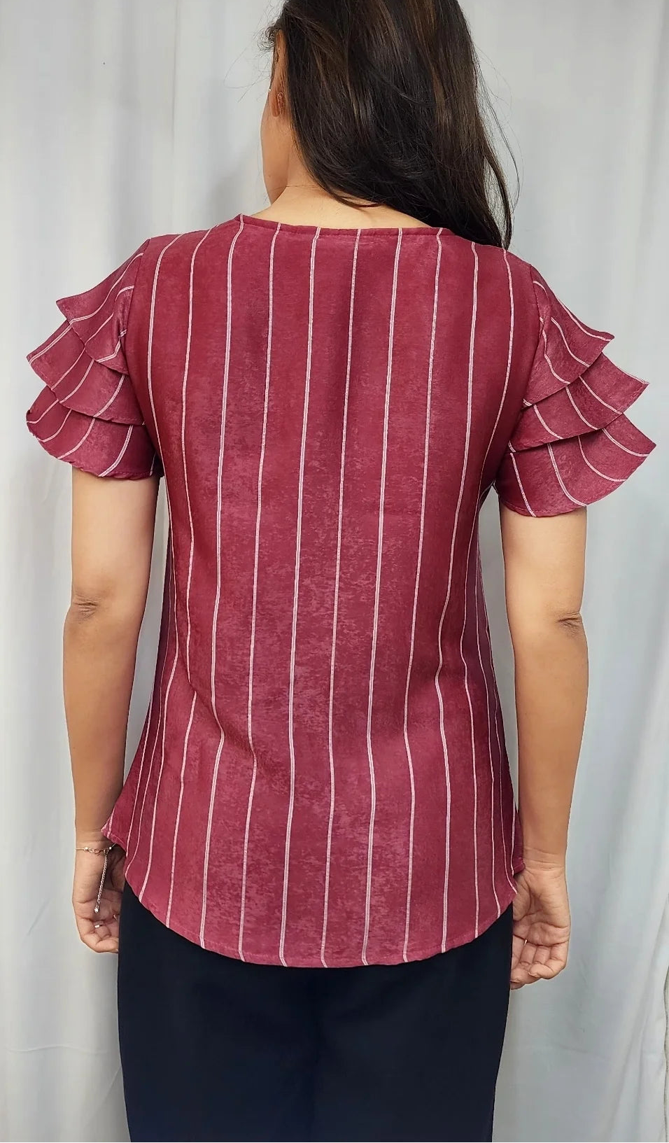 A woman viewed from the back wearing a dusty rose red short-sleeve top with thin white vertical stripes and layered ruffle details on the sleeves, paired with black pants.