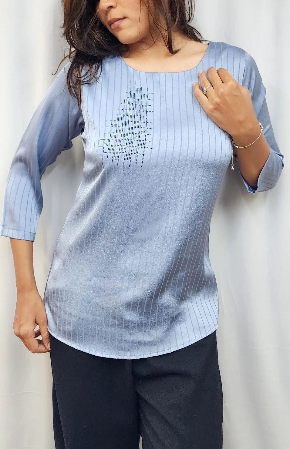 A woman wearing a light blue, three-quarter sleeve top with subtle vertical stripes and a geometric pattern on the chest, paired with dark pants, standing against a light-colored curtain.