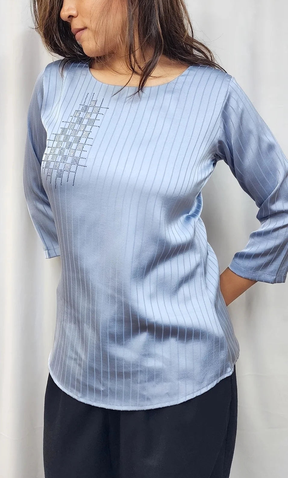 A woman wearing a light blue three-quarter sleeve top with subtle vertical stripes and a geometric pattern on the chest, paired with dark pants, set against a neutral light gray background.