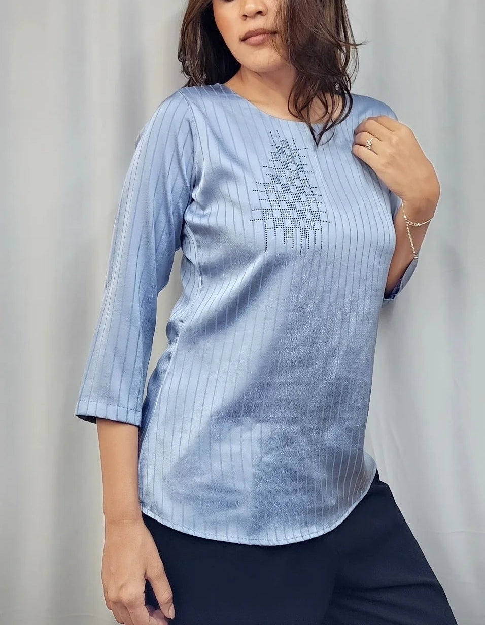 A woman modeling a light blue striped three-quarter sleeve top with a geometric embroidered pattern on the chest, paired with dark pants, standing in front of a white curtain.