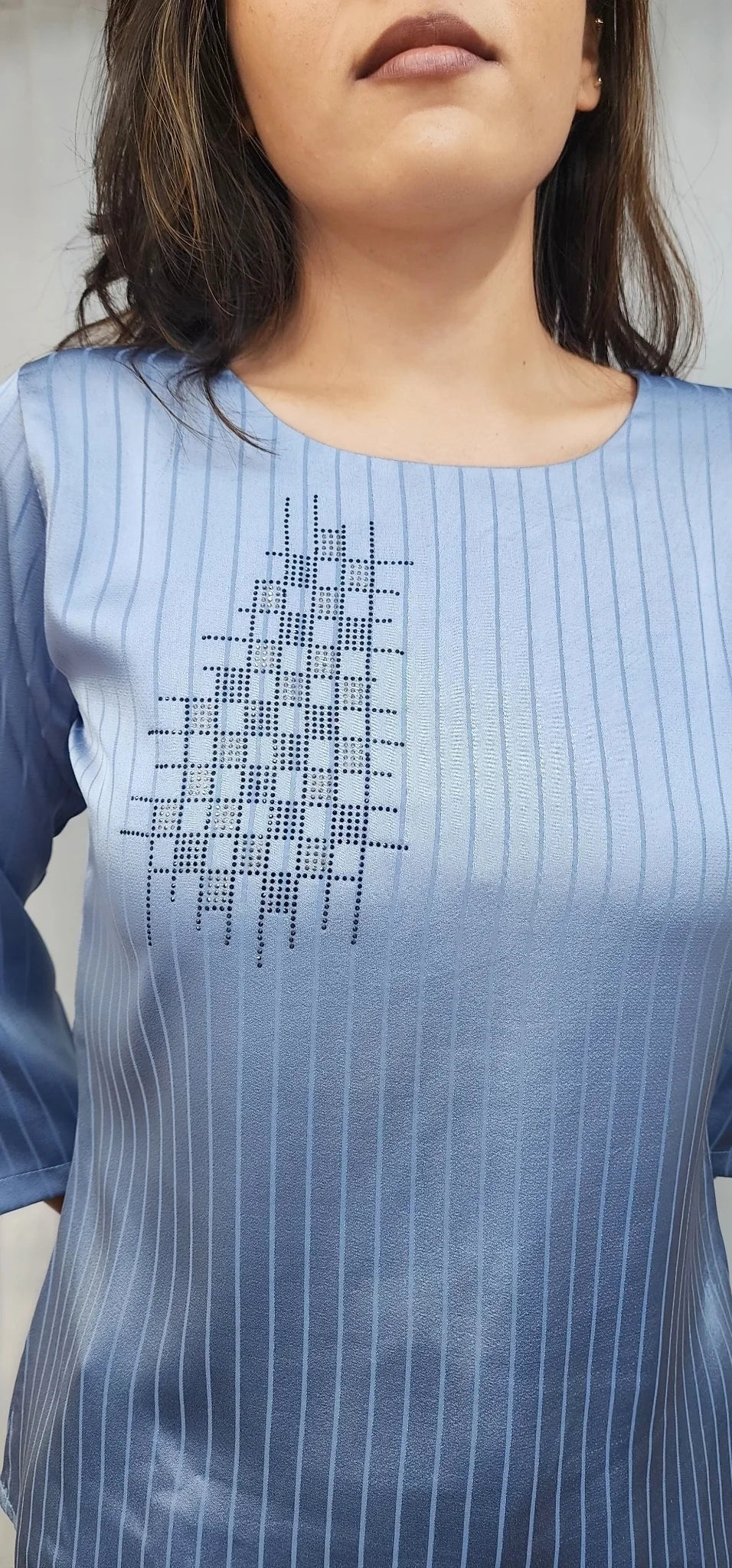 A close-up of a woman wearing a light blue pinstriped tunic top with a geometric embroidered pattern on the chest.