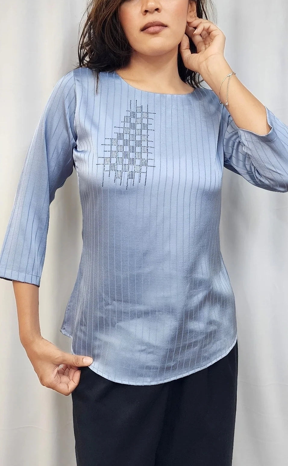 A woman modeling a light blue three-quarter sleeve top with a subtle vertical stripe pattern and a geometric embroidered design on the chest, paired with dark pants, set against a neutral draped backdrop.