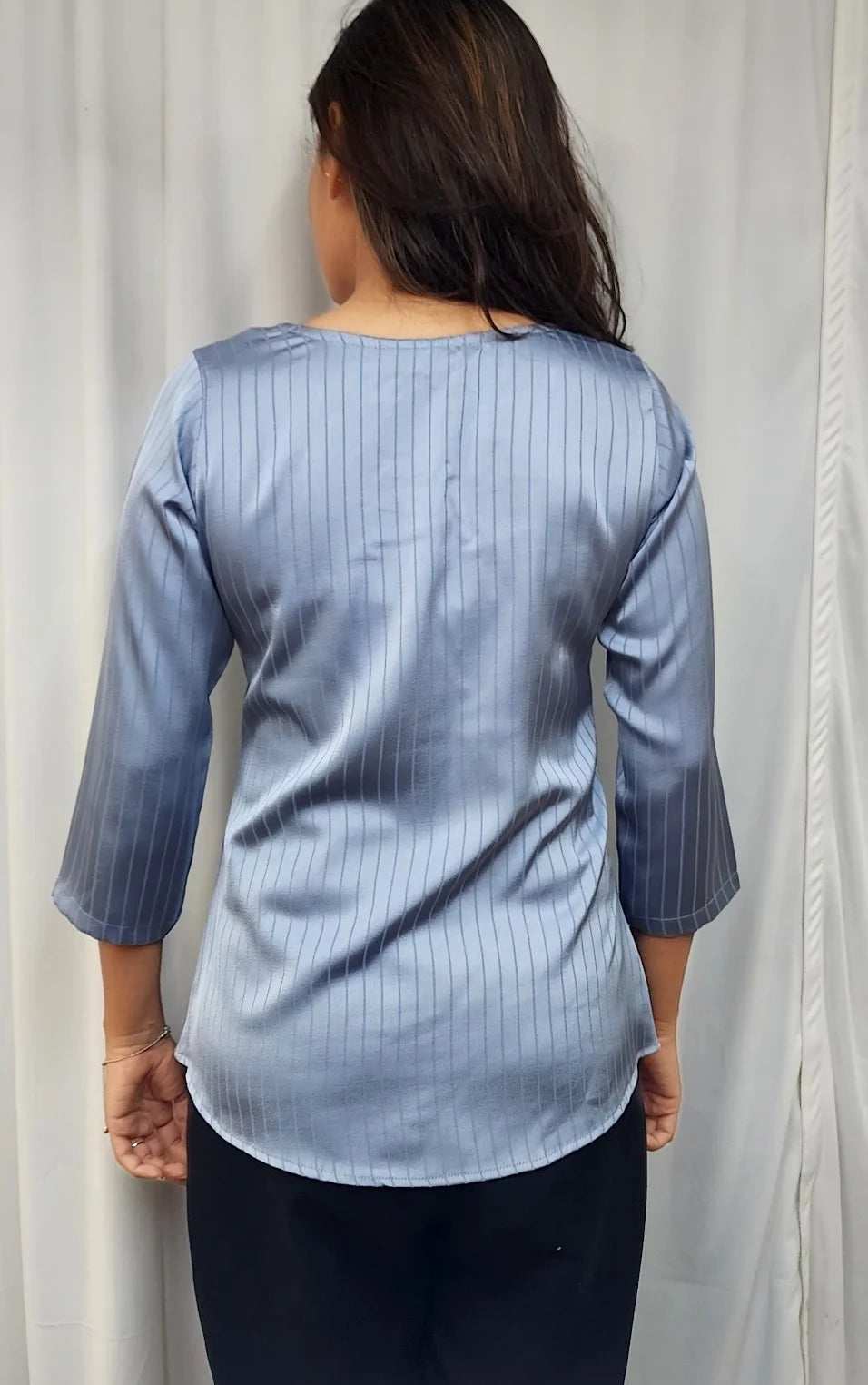 A woman viewed from behind wearing a light blue pinstriped blouse with three-quarter sleeves and black pants, standing in front of a white curtain.